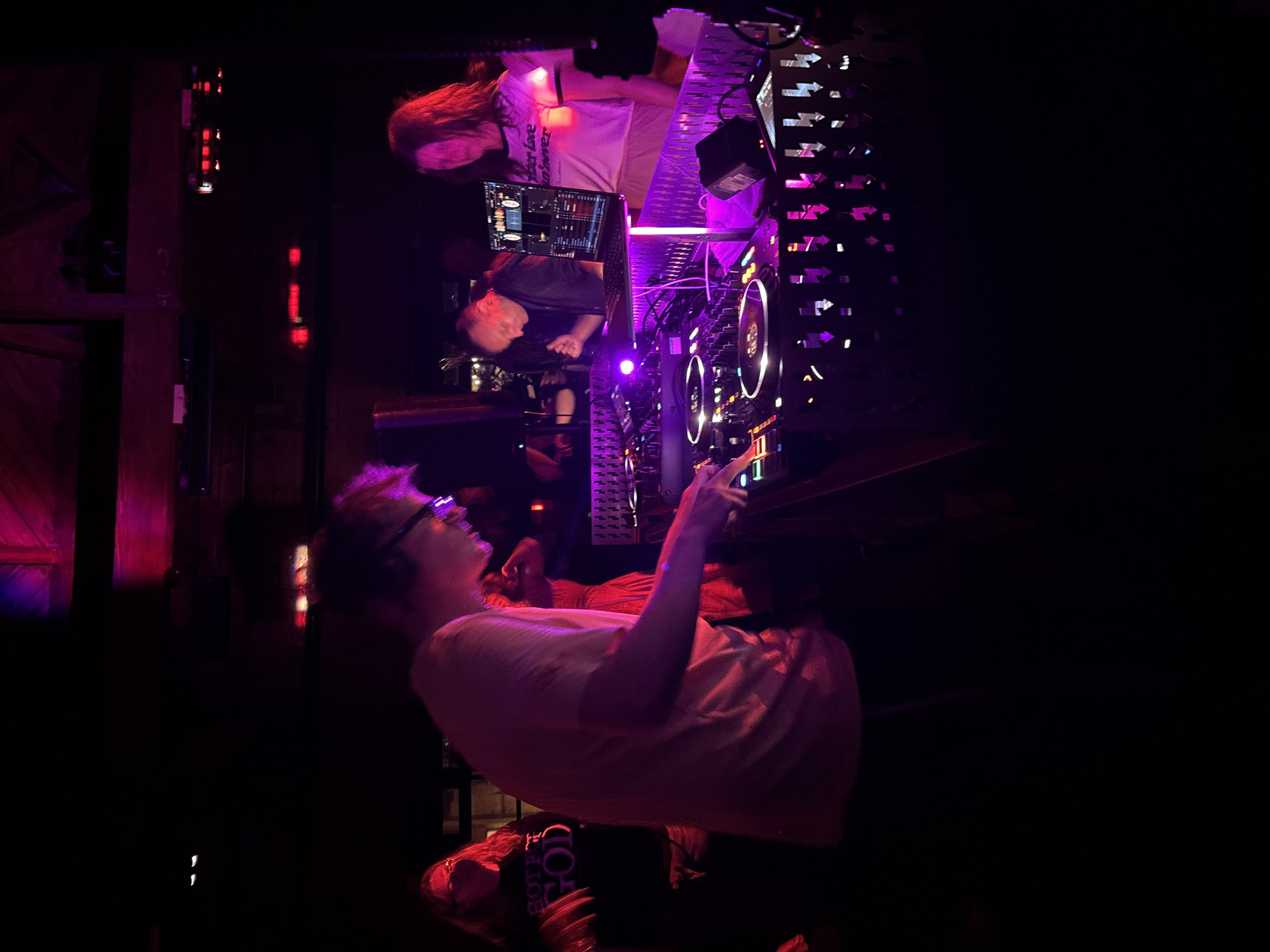 The image shows the interior of what appears to be a nightclub or music venue. A DJ is operating a mixing console, while another person stands nearby observing. The lighting is dark, with pink and purple hues illuminating the DJ equipment and surrounding area. The city is not identifiable in the image.