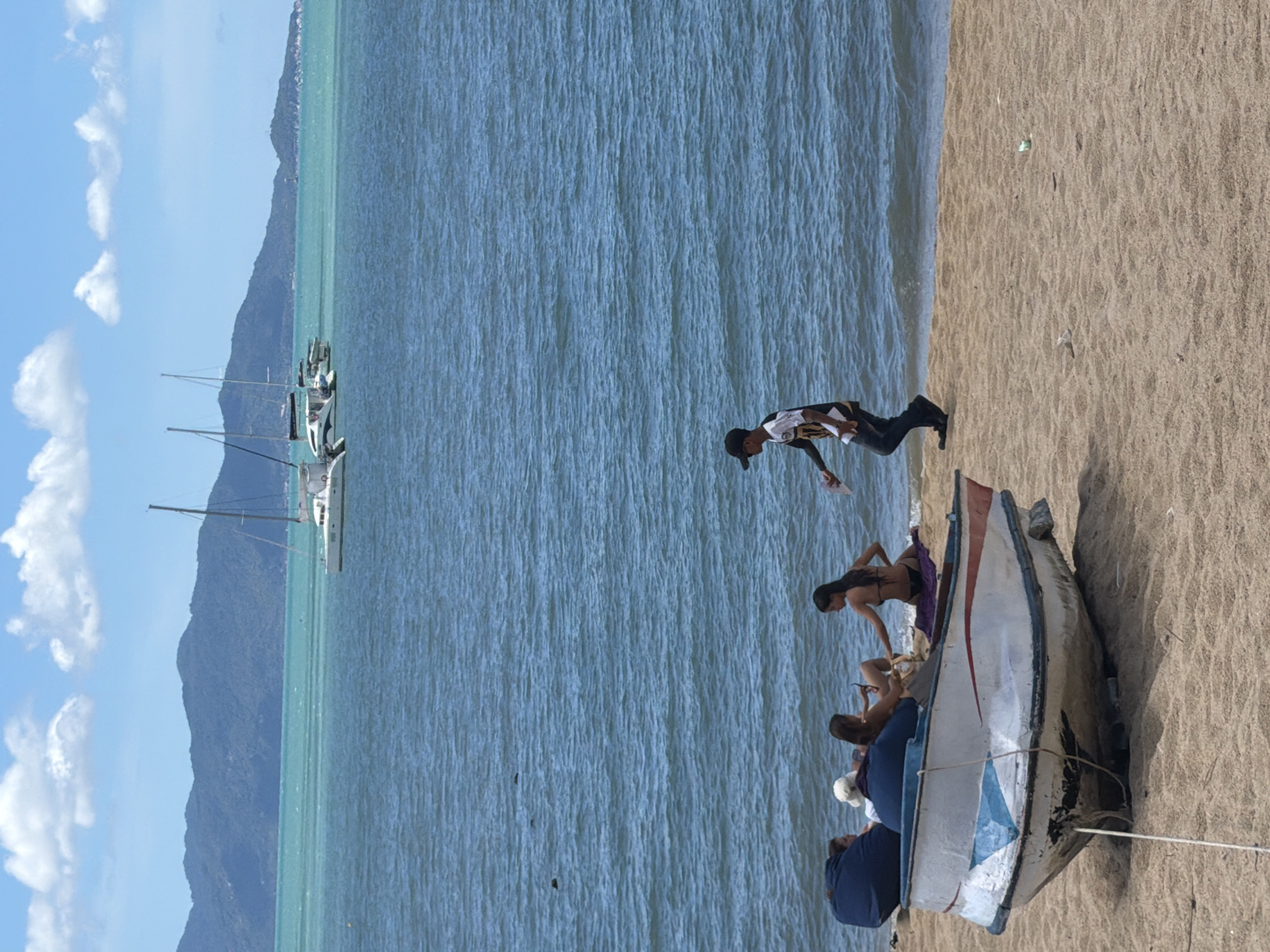 The image depicts a beach scene in Baan Bo Phut, Thailand. In the foreground, a small, weathered boat rests on the sand, partially occupied by four individuals. Three women, one wearing a white hat and another in a black bikini, are seated in the boat. A fourth woman, also in a bikini, is sitting on the sand next to the boat. A man wearing a black baseball cap and a patterned t-shirt is walking away from the boat towards the water. The ocean is calm with blue-green water, and several sailboats are visible in the distance. Behind the sailboats, a range of green mountains forms the horizon. The sky is blue with scattered white clouds. The sand is light brown.