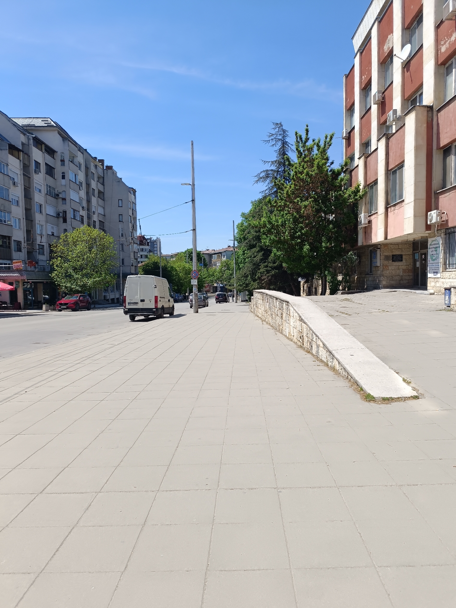 This is a daytime street scene in Pleven, Bulgaria, under a clear blue sky. The image captures a paved street with vehicles and sidewalks flanked by buildings.
On the left, a multi-story apartment building with numerous windows, balconies, and air conditioning units lines the road. A red car is parked in front of it, and a white van is driving away from the viewer on the road. Further down the street, more buildings and trees are visible.
On the right side of the image, another multi-story building stands, characterized by reddish-brown and lighter vertical panels, many windows, and several exterior air conditioning units and a satellite dish. This building shows signs of wear on its facade. In front of this building, a concrete ramp with a low stone wall leads up from the main sidewalk. Adjacent to the building's entrance, there is a sign in Cyrillic that reads "РЕГИОННО КРЪВОДАРИТЕЛСКИ ЦЕНТЪР" (Regional Blood Donation Center) and indicates working hours "07:30 - 13:00". A tall street light pole with a 'No Stopping' sign is positioned near the road. Various green trees are scattered throughout the scene, adding greenery. The sidewalk in the foreground is composed of light-colored square paving tiles.
The weather is sunny and clear, suggesting a pleasant day. No people are visible, depicting an ordinary moment in an urban environment.