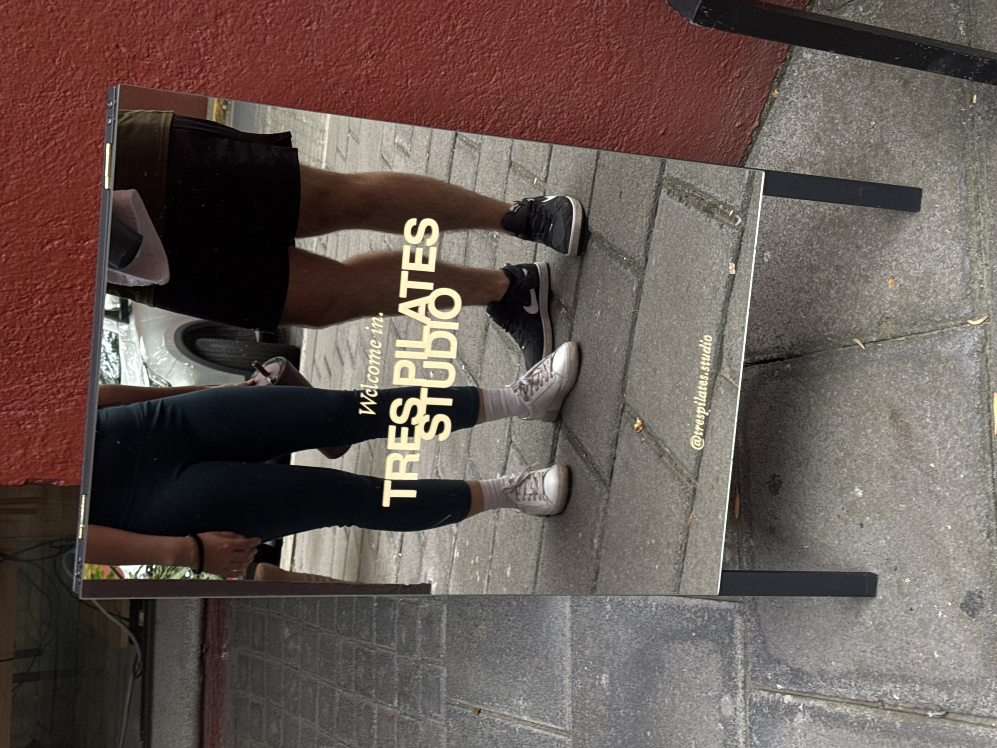 The image shows a mirrored sandwich board sign for "Tres Pilates Studio" in Puebla, Mexico. The sign's mirrored surface reflects the streetscape, including two pedestrians. One pedestrian, presumably male, is wearing black shorts and sneakers, while the other, likely female, is wearing dark leggings and white sneakers. The sign is placed on a sidewalk next to a red wall and gray pavement. The sign also shows a reflection of a parked vehicle.