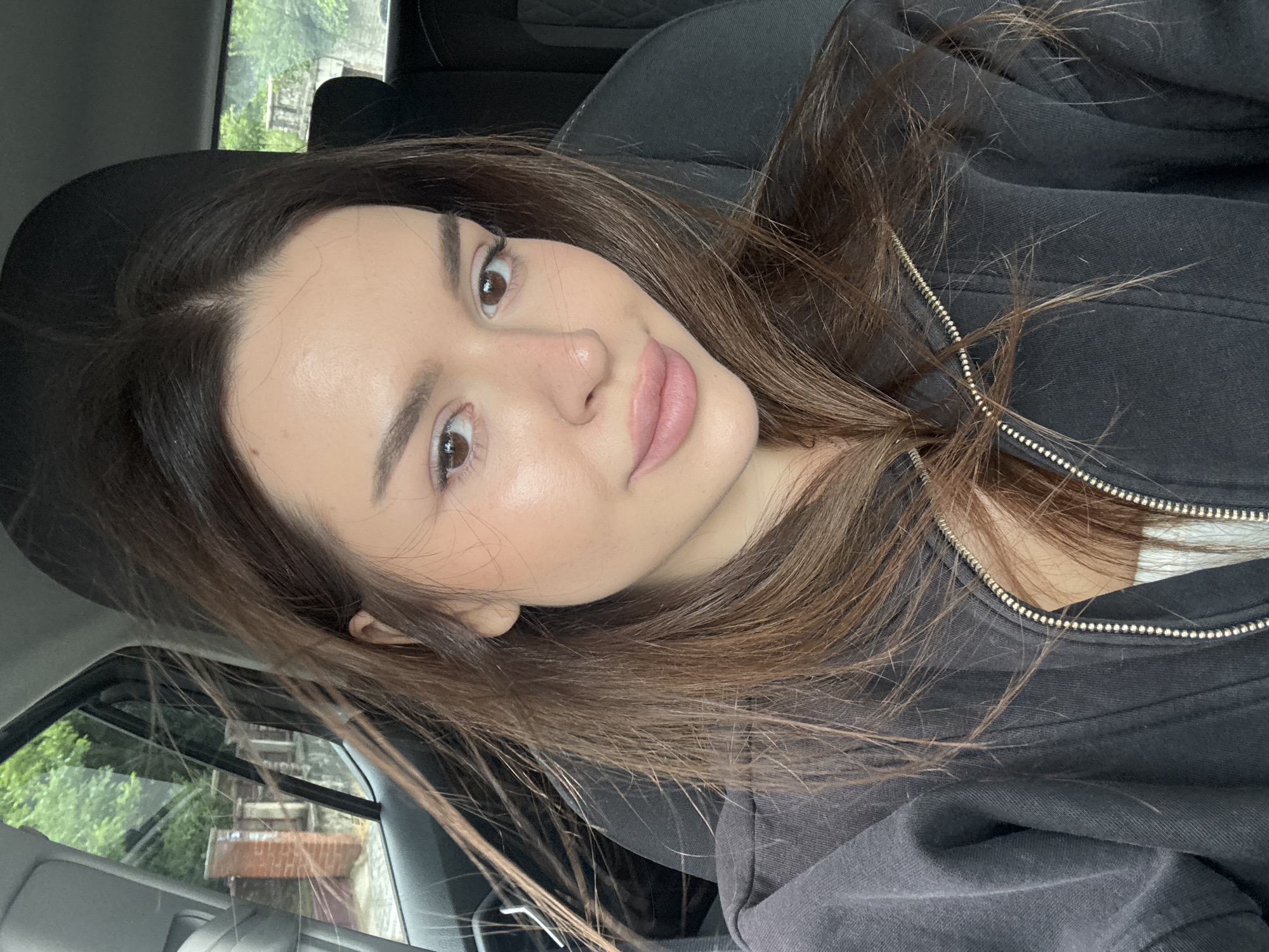 The media file is a close-up, vertically oriented selfie of a young woman inside a car, likely in Prague, Czechia. She has long, brown hair, brown eyes, and fair skin with light makeup. Her lips are slightly parted, and she is looking towards the camera with a gentle, relaxed expression. She is wearing a black zip-up hoodie.
The background shows the interior of a car, with a dark headrest visible. Through the car window to the left, there is a glimpse of an outdoor scene with greenery, suggesting it is daytime. The light appears soft, possibly indicating overcast weather or the time of day being early morning or late afternoon. The exterior scenery, visible through the window, shows some architectural elements and trees.
The overall impression is that of a casual, candid moment captured while traveling in a vehicle. There is no visible text in the image.