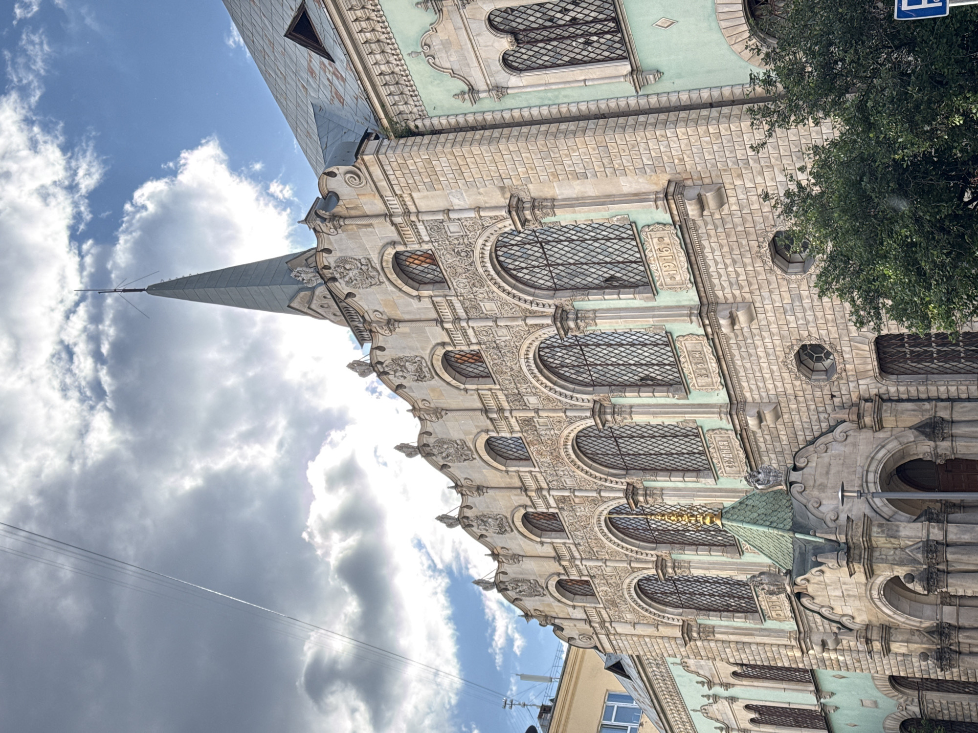 A low-angle shot shows a section of a richly decorated building against a sky with clouds. The building features rows of arched windows with intricate metal grilles. Above the windows, there are detailed carvings and decorative elements. The building has a pointed spire rising into the sky. The architecture is ornate and appears to be of historical significance. The scene is well-lit. Moscow, Russia is not identifiable in the image.