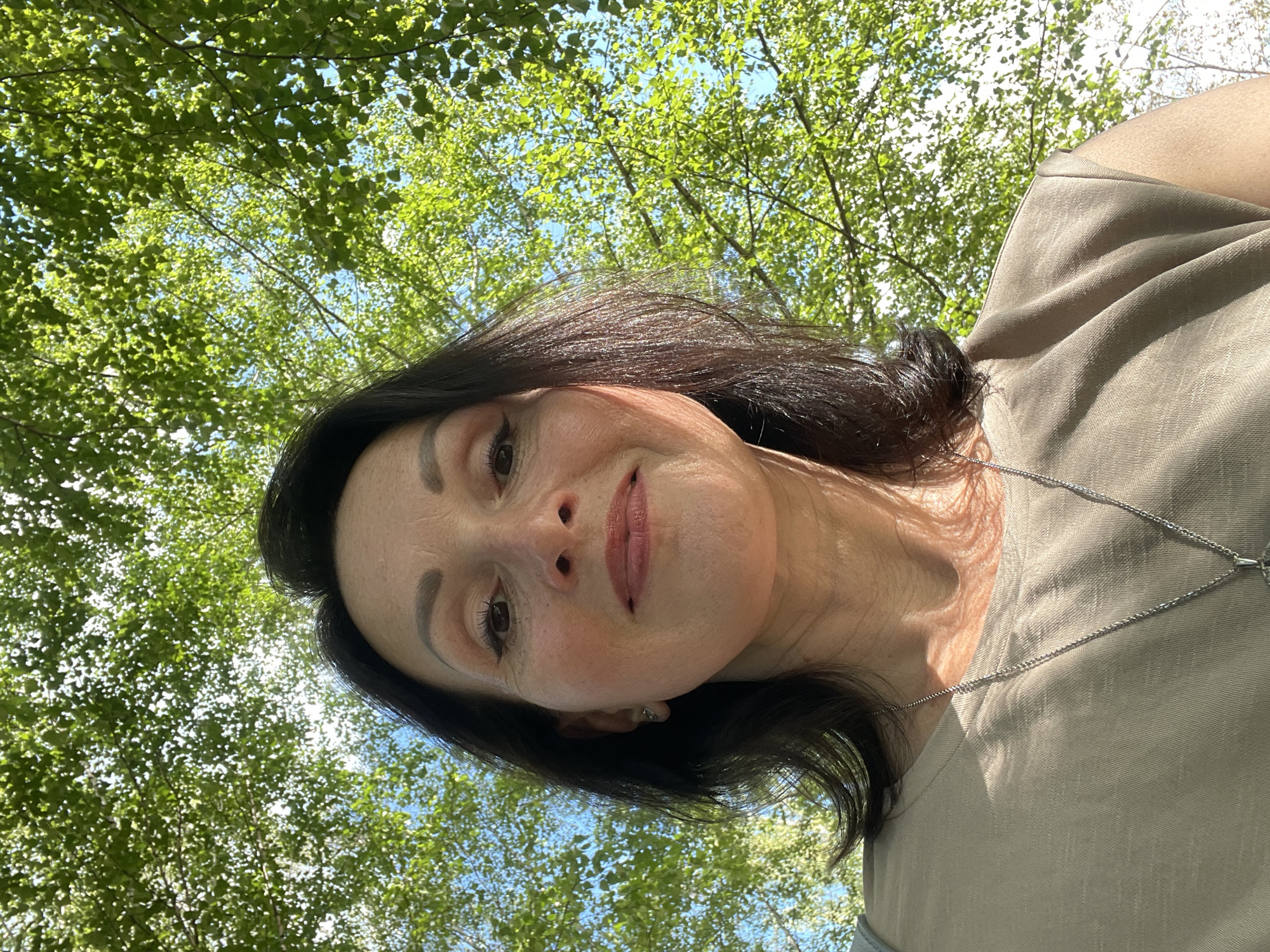 A woman with dark hair, wearing a beige t-shirt and a silver necklace, looks up at the camera from a low angle. She has makeup on, including eyeliner and lipstick. Her face is illuminated by sunlight, casting shadows on her skin. Behind her, green leaves and blue sky are visible, suggesting an outdoor setting, possibly under a tree. The image does not provide any specific details that would allow for identification of the city of Kirovo-Chepetsk, Russia.