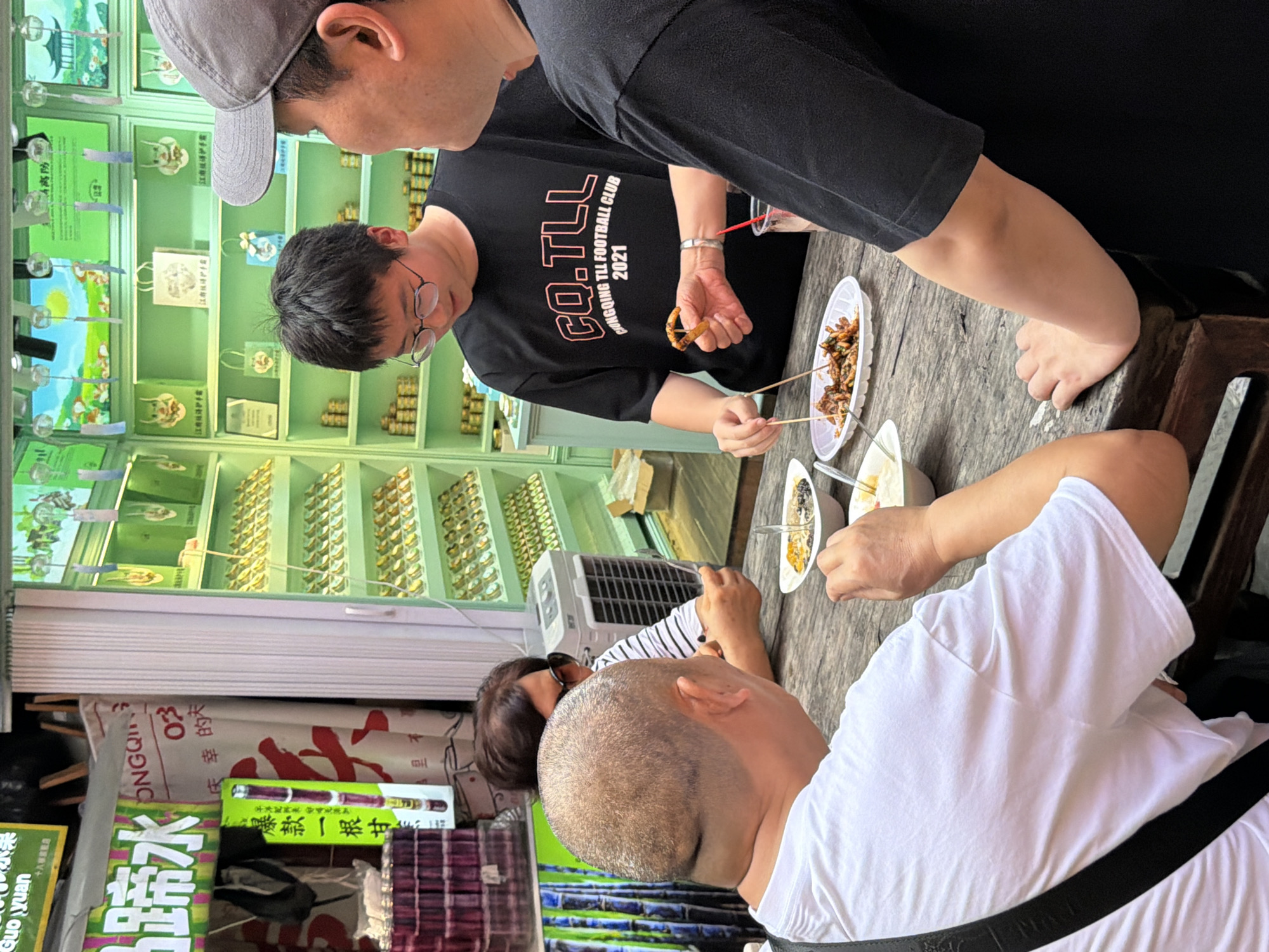 In a casual outdoor setting, likely a street-side eatery or market stall in Yuzhong District, China, three individuals are gathered around a wooden table. The lighting suggests daytime. Two men and one woman are present. One man, wearing a grey baseball cap and a black t-shirt with "CO.TLL CJONGQING TLL FOOTBALL CLUB 2021" printed on it, is reaching for food with skewers from a white plastic container. Another man, with a shaved head, is seated and facing away from the camera, wearing a white t-shirt. The woman, wearing glasses and a striped shirt, is also seated at the table. In the background, shelves are filled with various products, appearing to be health and beauty items, possibly skincare, with packaging featuring Chinese text such as "江护手霜" (Jiang Hu Shou Shuang - Jiang protection hand cream) and "庆幸的天" (Qing Xing De Tian - Blessed day). Several food items are visible on the table, including what appears to be fried snacks on skewers and dips in small bowls. Signs with Chinese characters are also visible, including "蹄水" (Ti Shui - Hoof water, likely a brand name or product), "ONGQI", "Guo yuan", and "爆款一根甘" (Bao Kuan Yi Gen Gan - Hot style one sweet). The overall scene depicts a group of people enjoying a meal and possibly shopping for goods.