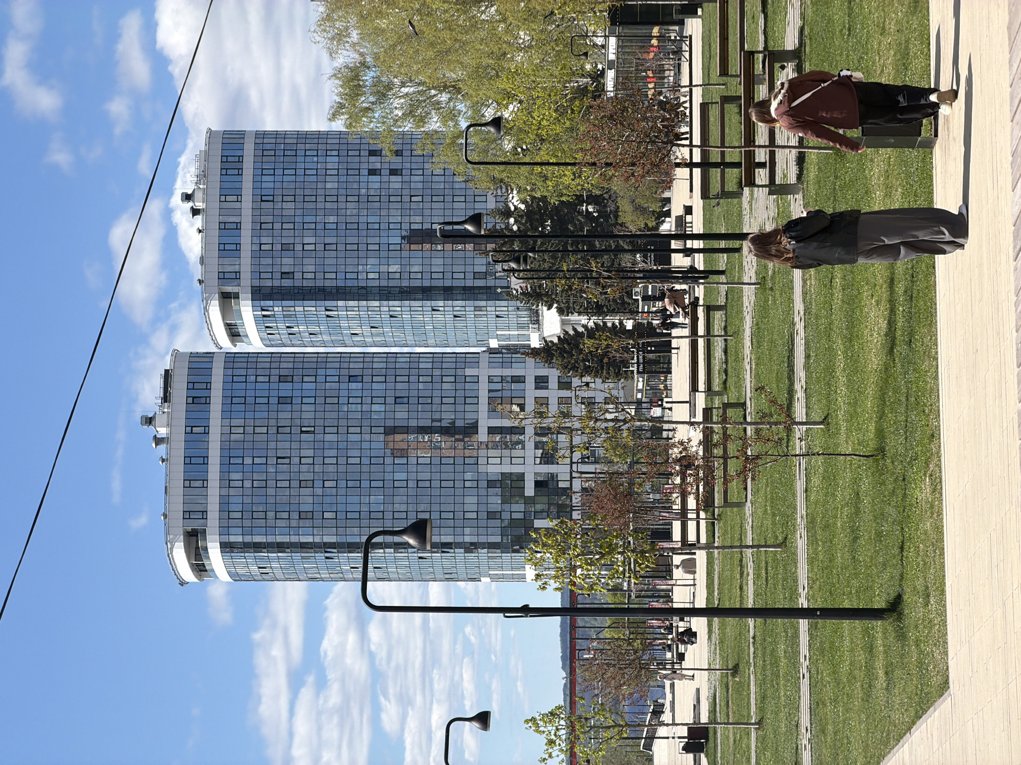 The image shows an outdoor scene in Izhevsk, Russia, featuring two modern, cylindrical-shaped apartment buildings with glass facades under a blue sky with scattered clouds. A power line is visible on the left. The scene is taken from a slightly elevated perspective.
In the foreground, there is a grassy park area lined with black lampposts and small trees. Two individuals are present: one wearing a grey jacket and standing while facing away from the camera, and another wearing a reddish-brown jacket sitting on a park bench. A beige paved area is located on the right side. The image is taken on a clear, sunny day.
