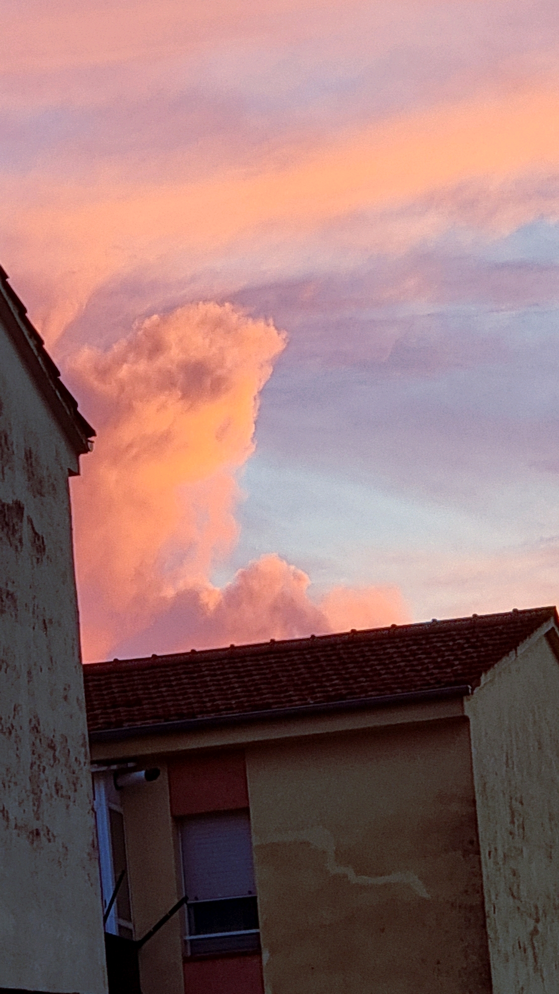 A photograph captures a sunset over Sant Joan Les Fonts, Spain, with dramatic clouds painted in hues of pink, orange, and purple. The sky dominates the upper portion of the image, transitioning from soft pastels to deeper, richer tones. A prominent, large cumulus cloud formation, illuminated by the setting sun, takes center stage in the middle of the sky. Below the sky, the tops of buildings are visible, silhouetted against the vibrant sky. Two buildings are partially captured in the frame: one on the left, a lighter-colored structure with a weathered texture, and another on the right, featuring a terracotta-tiled roof. The building on the right also shows a section of a yellow facade with a window. The scene is static, with no visible people or immediate actions. The environment suggests an urban or suburban setting within Sant Joan Les Fonts, characterized by residential buildings against a backdrop of a breathtaking sunset.