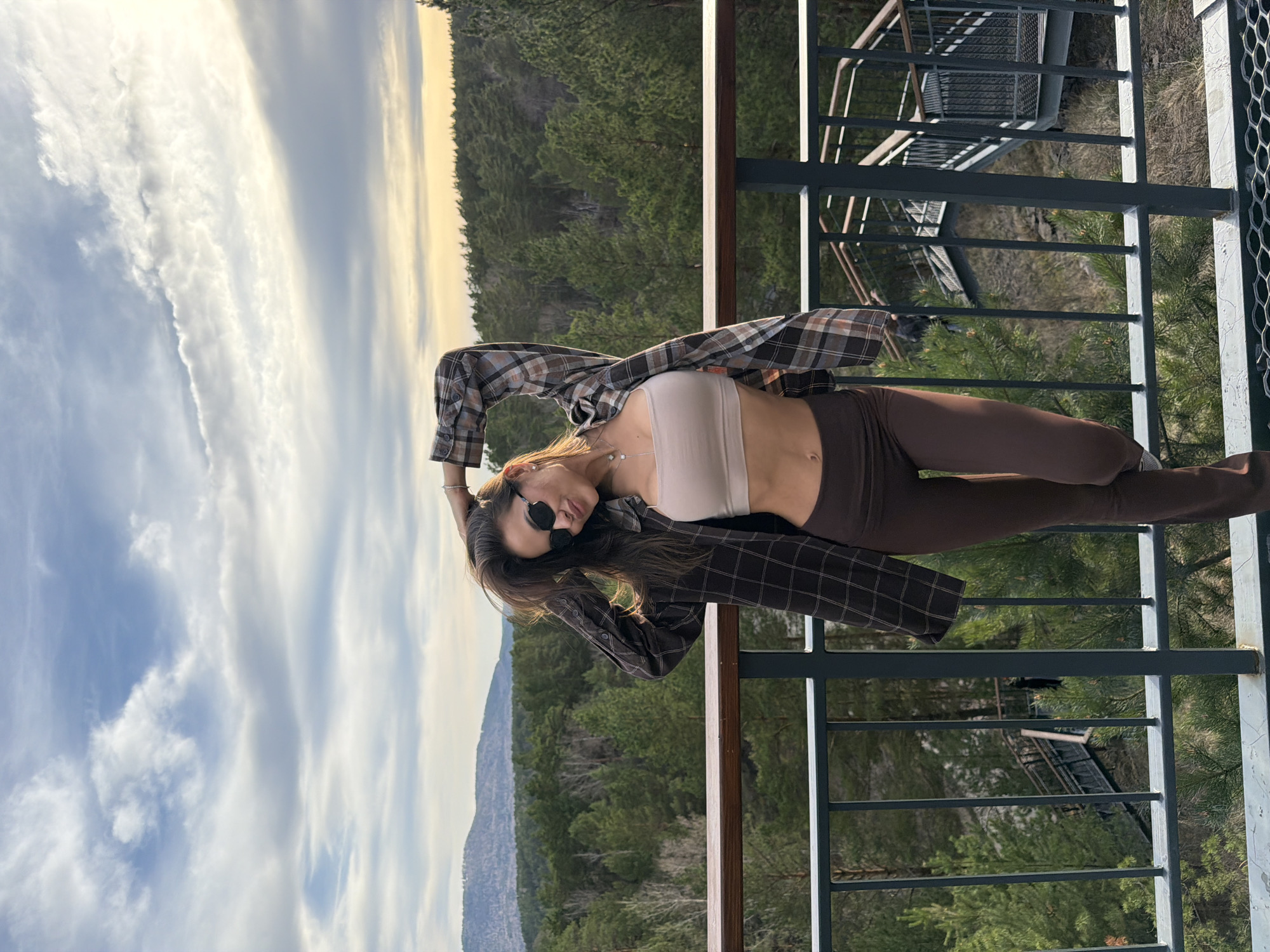 A slender woman is angled back with her head towards the left side of the frame, wearing a light-colored bandeau top, dark brown leggings, a dark plaid open shirt, and sunglasses. She is standing on a metal balcony, which is in focus. In the background, out of focus, there is a forest, a mountainous landscape, and a cloudy sky. This location might be in Krasnoyarsk, Russia, given that the image was provided with that designation.