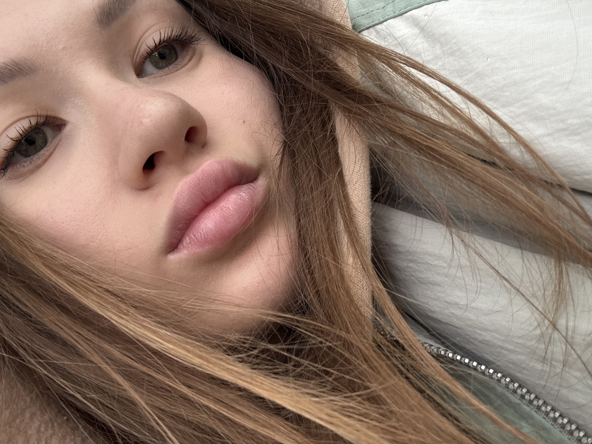 A close-up photograph features the face of a young woman with light brown hair. Her skin is fair, with subtle blemishes on her cheek. Her eyes are hazel and are lined with dark mascara. Her lips are full and tinted a light pink. Some strands of her hair fall across her face. She is wearing a hooded jacket that is light blue on the outside and light tan on the inside. The zipper of the jacket is visible. The location is unspecified and cannot be determined from the image.