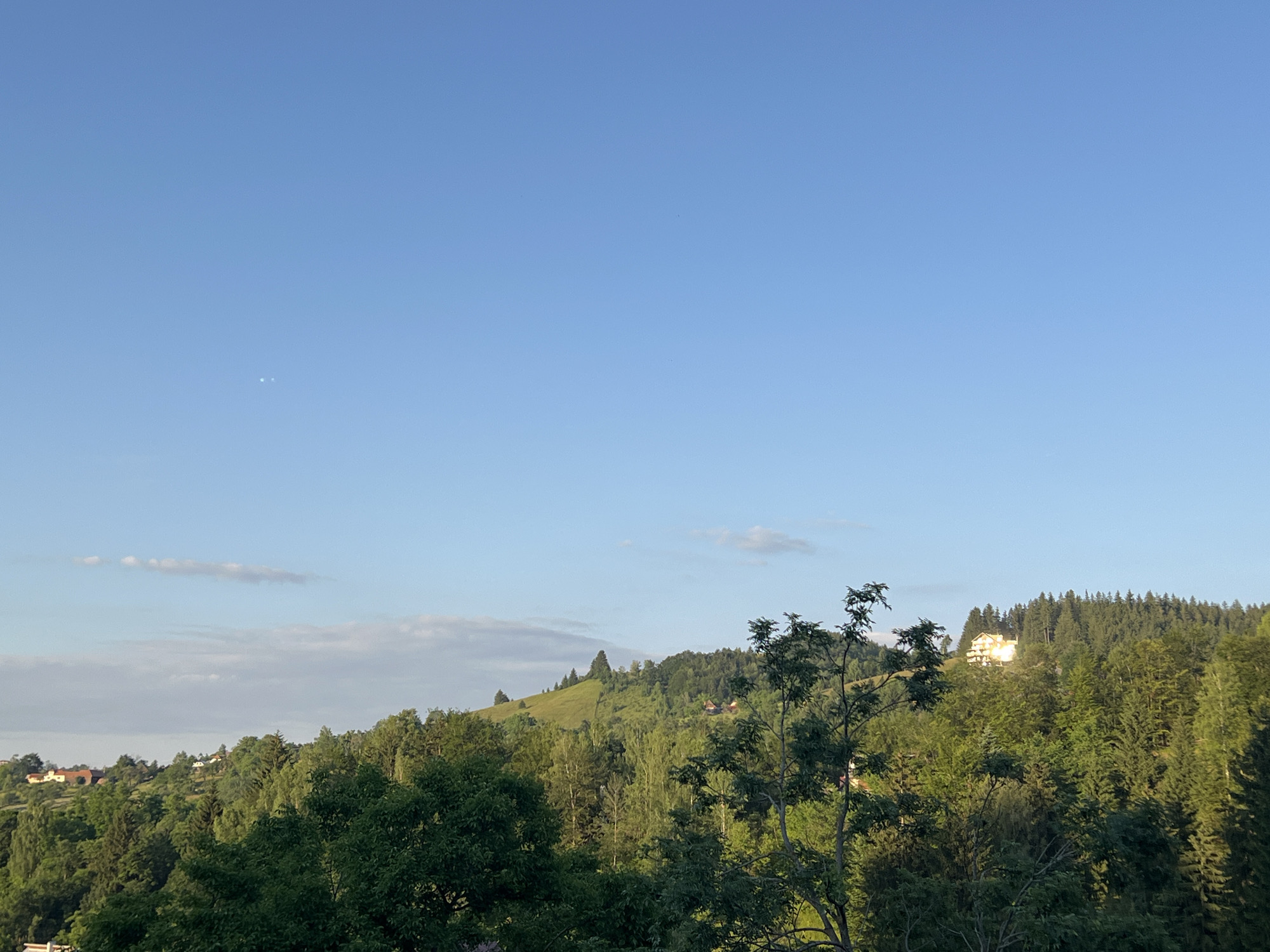 The image captures a serene, verdant landscape under a clear blue sky with a few wispy clouds. The scene is dominated by rolling hills covered in a dense forest of various shades of green. In the foreground, the lush foliage of trees forms a natural frame, with some branches reaching towards the sky.
Nestled on one of the hills, a bright white building with a red-tiled roof is visible, its windows reflecting the sunlight. Further in the distance, scattered houses with similar red roofs are interspersed among the trees on other hillsides, suggesting a rural or semi-rural setting. The sunlight illuminates the landscape, creating a warm and inviting atmosphere.
Two small, bright specks are faintly visible in the upper left quadrant of the sky, possibly celestial bodies. The overall impression is one of natural beauty and tranquility, with hints of human habitation integrated harmoniously into the environment. The time of day appears to be late afternoon or early evening, given the angle of the sunlight and the soft light illuminating the scene. The weather seems pleasant and clear. No text is visible in the image.