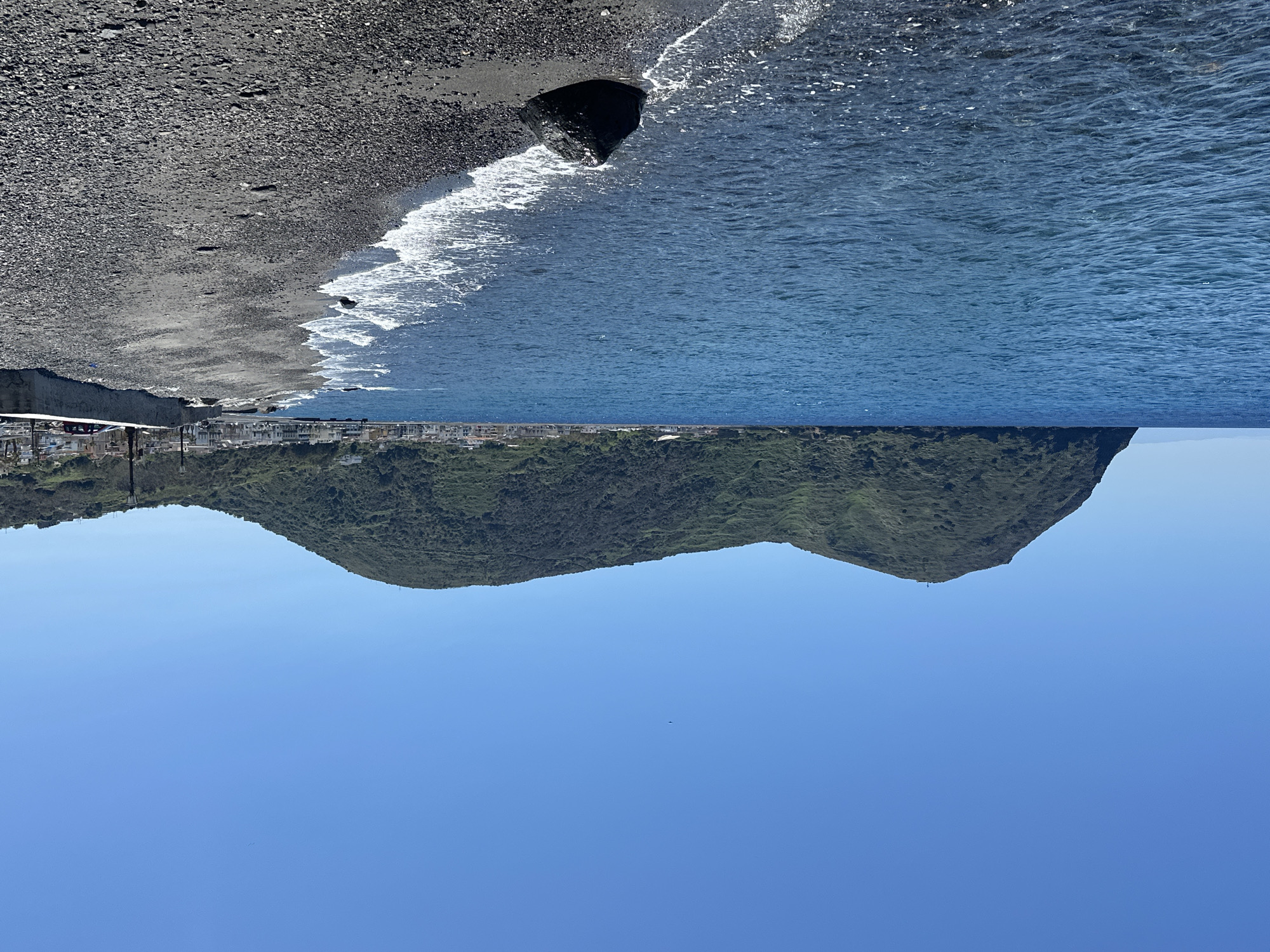 The image shows a coastal scene, likely in Lipari, Italy. The foreground features a rocky beach of dark, volcanic stones meeting the edge of the ocean, with gentle waves washing ashore. The middle ground displays a green, mountainous landmass with a small town visible at the base, which hints at a settlement along the coastline. The backdrop is dominated by a clear, blue sky with no visible clouds. The image appears to be inverted.