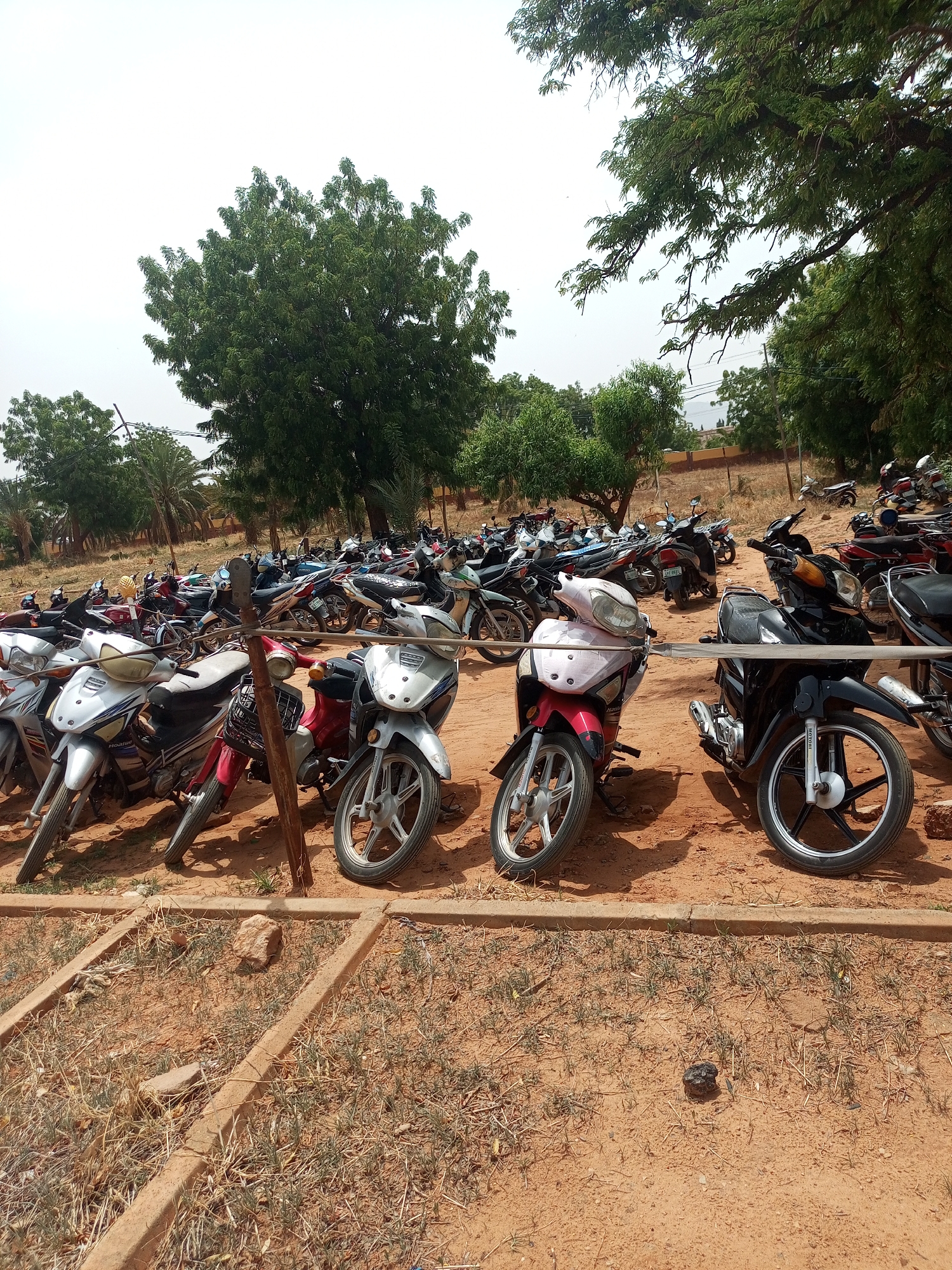 The image shows a parking area in Gombe, Nigeria, filled with numerous motorcycles. The ground is reddish-brown dirt with sparse vegetation. Concrete barriers define parking spaces in the foreground. The motorcycles vary in color and style. Trees provide shade. The sky is overcast.