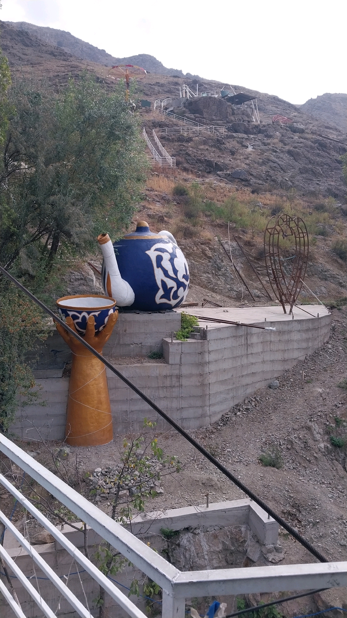 The image shows an outdoor scene in the city of Oltinkon, Uzbekistan, featuring large decorative objects on a hillside. In the foreground, a large blue and white teapot sculpture is prominently displayed on a concrete platform. To the left of the teapot is another sculpture resembling a golden hand holding a patterned bowl. The hillside is rocky and sparsely vegetated, with a few trees visible. In the background, a fairground ride with colorful umbrellas is situated further up the hill, and white railings line a series of stairs leading up the slope. A thick cable runs diagonally across the image, likely part of a cable car system given the mountainous terrain. The sky is overcast.