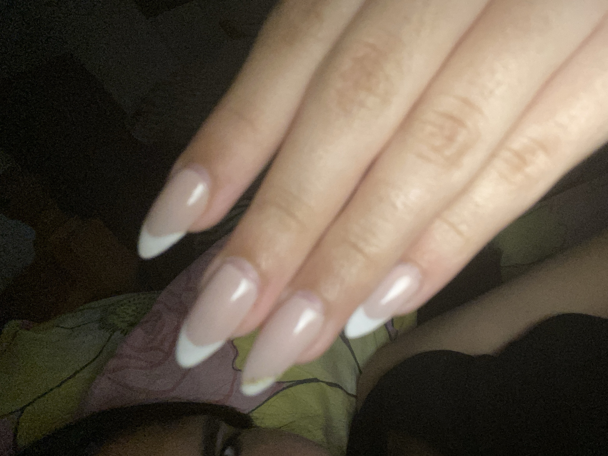 A close-up photograph depicts a person's hand with almond-shaped nails that have a French manicure design. The nails are a pale, neutral color with white tips. The hand is positioned in front of a background that includes a patterned fabric, possibly a bedsheet, and a dark garment. Part of a face is visible in the lower left corner. The setting is dark, with low ambient light. There is no evidence to indicate this photo was taken in Cotoperíz, Venezuela.