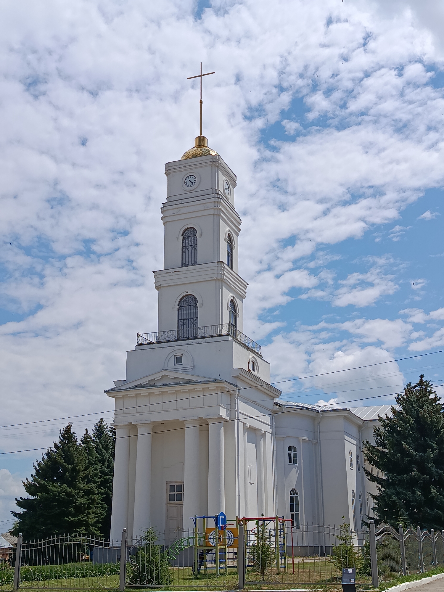 A white church with a tall, square bell tower topped with a golden dome and cross stands against a bright blue sky filled with fluffy white clouds. The bell tower has clocks on its upper section and arched windows with decorative metal grates. A portico with large white columns supports the lower part of the tower. To the right of the bell tower, the white church building extends with arched windows. Evergreen trees are visible behind the church. In the foreground, a decorative metal fence encloses a grassy area with a small children's playground visible behind it. The scene is set in the city of Marx, Russia.