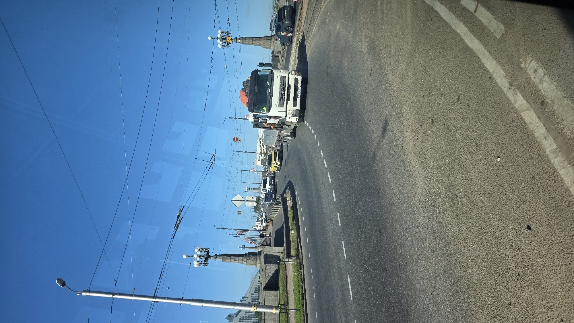 The image shows a view of the Akmens Bridge in Riga, Latvia, on a clear, sunny day. The bridge spans the Daugava River. A white flatbed truck with an orange crane is driving across the bridge. Other vehicles, including a yellow car and a white van, are also visible on the road. Ornate streetlights line the bridge. In the background, a modern building and a Ferris wheel can be seen. The road surface is asphalt with dashed white lane markings. Overhead, power lines and trolleybus wires crisscross the sky.