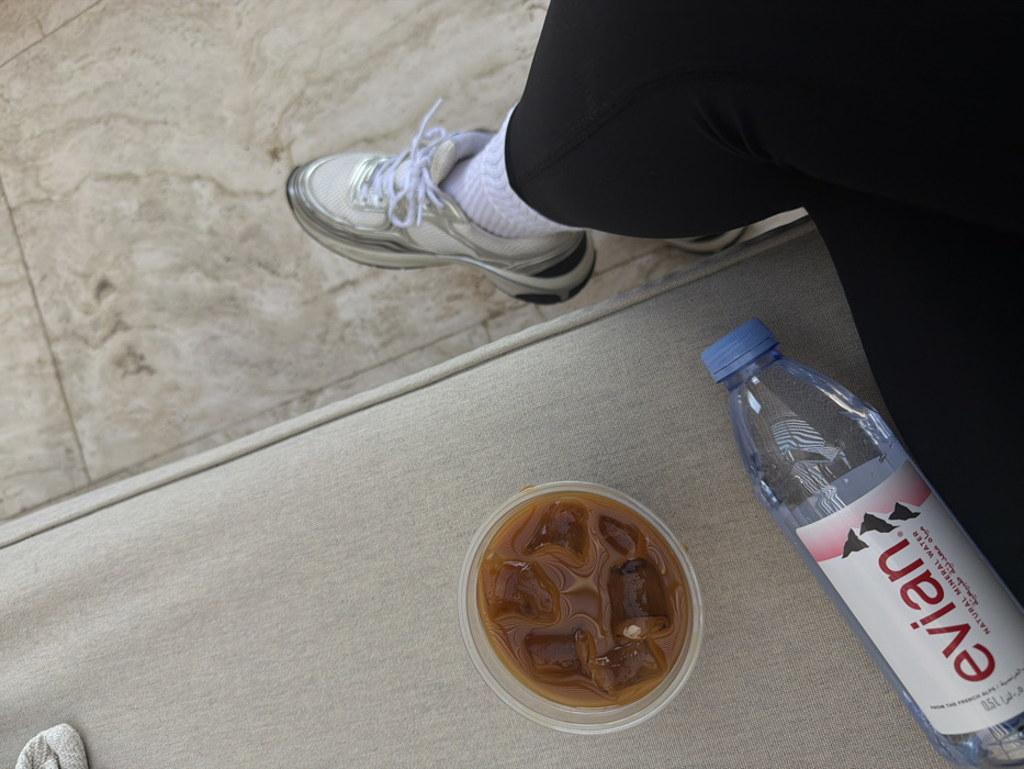 An overhead view captures the lower right leg of an individual, clad in black athletic leggings, a white ribbed sock, and a white and silver athletic shoe. Adjacent to the leg, on a light beige fabric-covered surface, rests a clear plastic cup containing an iced dark brown beverage with multiple ice cubes. Next to the cup, a 0.5L Evian natural mineral water bottle with a blue screw cap is visible. The individual's right foot, encased in the athletic shoe, is positioned on a light-colored, patterned tiled floor. The scene is located in Dubai, United Arab Emirates.
