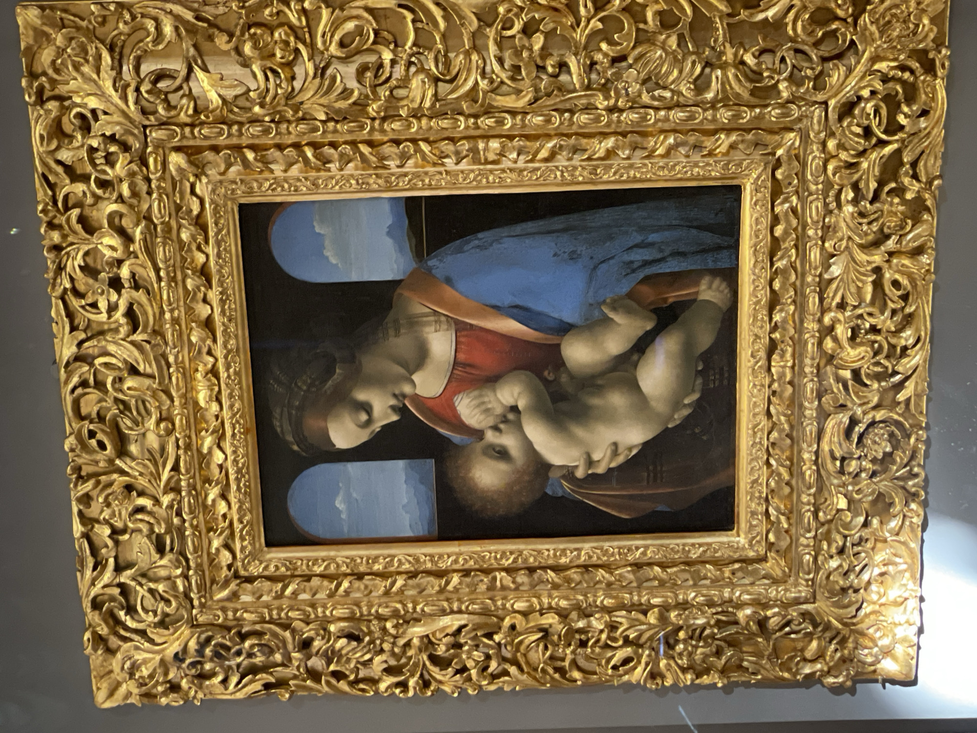 The image shows a painting, possibly "Madonna and Child," with a large, ornate gold frame. The painting depicts a woman in a red dress with a blue draped around her, cradling an infant. Two arched windows are visible in the background, showing a sky. The painting appears to be hanging on a wall, likely in a museum or gallery in the city of Unknown, Unknown.