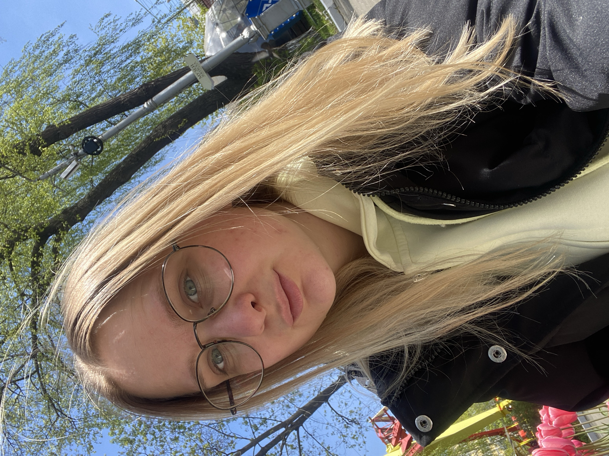 A selfie-style, angled photograph shows a young, fair-skinned woman with long blonde hair and round spectacles. She wears a light-colored hoodie under a dark jacket. The background features a blue sky, bare tree branches, and an urban environment with what appears to be streetlights. The image suggests an outdoor location, possibly in a city park or street. Without additional details, it is impossible to confirm it's Izhevsk, Russia.