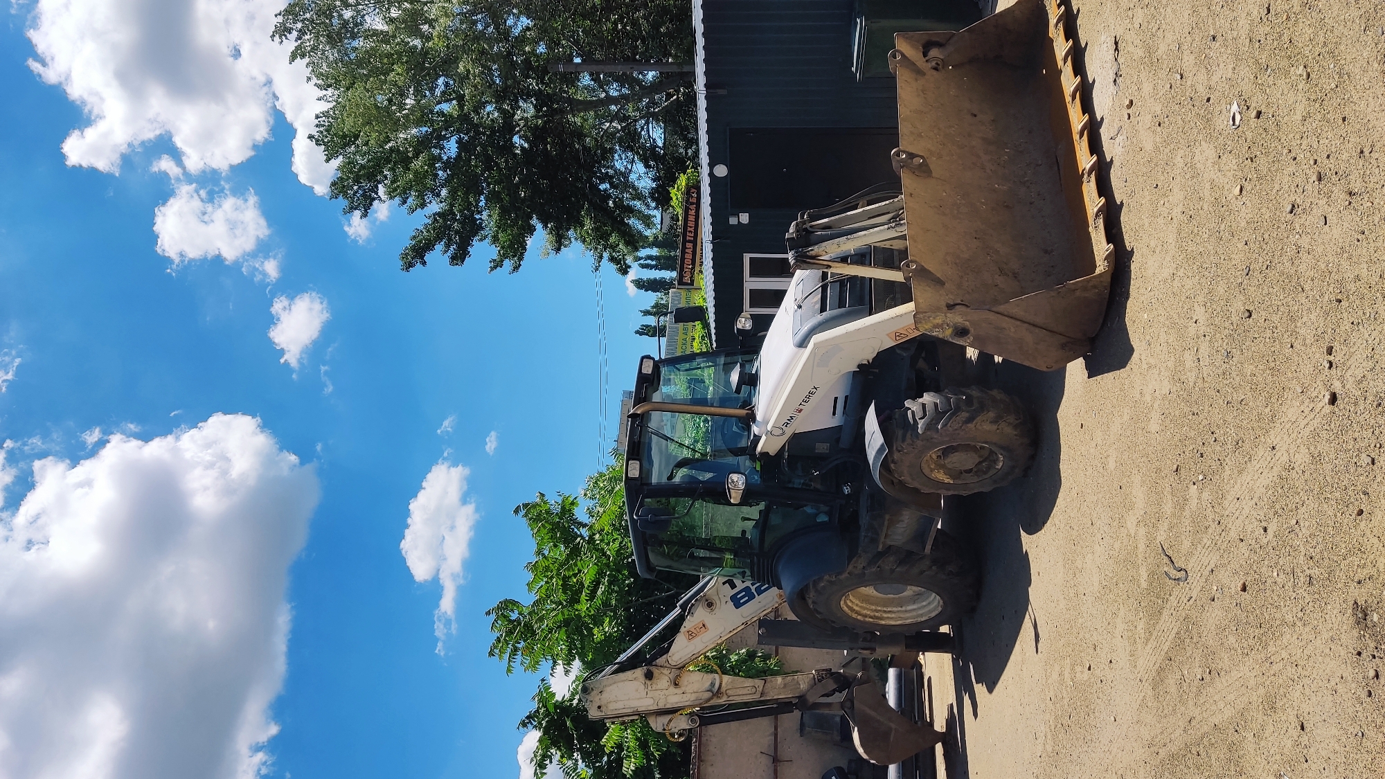 An outdoor, ground-level, medium shot captures a white and dark gray backhoe loader parked on a dirt surface under a bright blue sky with scattered white clouds. The loader is positioned in the center of the frame, facing slightly to the right. Its front loader is down and extended, and its rear digging arm is raised and angled. A partially visible building with a dark green corrugated metal roof and a white-trimmed window is situated behind the loader to the right. To the left of the loader, a light-colored wall and some pipes are visible. Trees and foliage form a backdrop behind the building and wall. A sign with Cyrillic text, "РЕМОНТ ТЕХНИКИ" (Repair of Equipment), is mounted on the building. The overall scene appears to be an industrial or service area. The presence of construction equipment and the sign suggest this location may be a repair shop or storage yard for heavy machinery in Krasnodar, Russia.