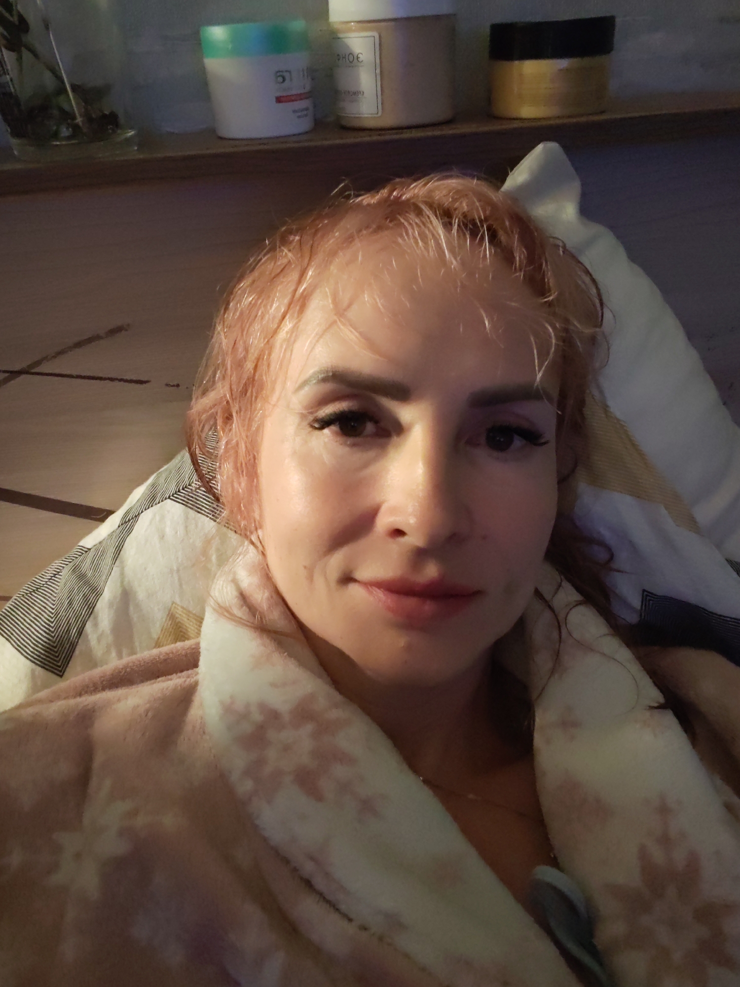 A woman with reddish-pink hair is lying on a bed, looking at the camera. She is wearing a light pink bathrobe with a snowflake pattern. Her hair is damp, and she appears to have makeup on, including false eyelashes and eyeshadow. She is lying on a pillow and a comforter with a striped and geometric pattern. In the background, on a shelf, are several jars and containers, possibly toiletries or cosmetics. The setting appears to be indoors, likely a bedroom. There is no visible indication of the specific location within Chaikovsky, Russia.