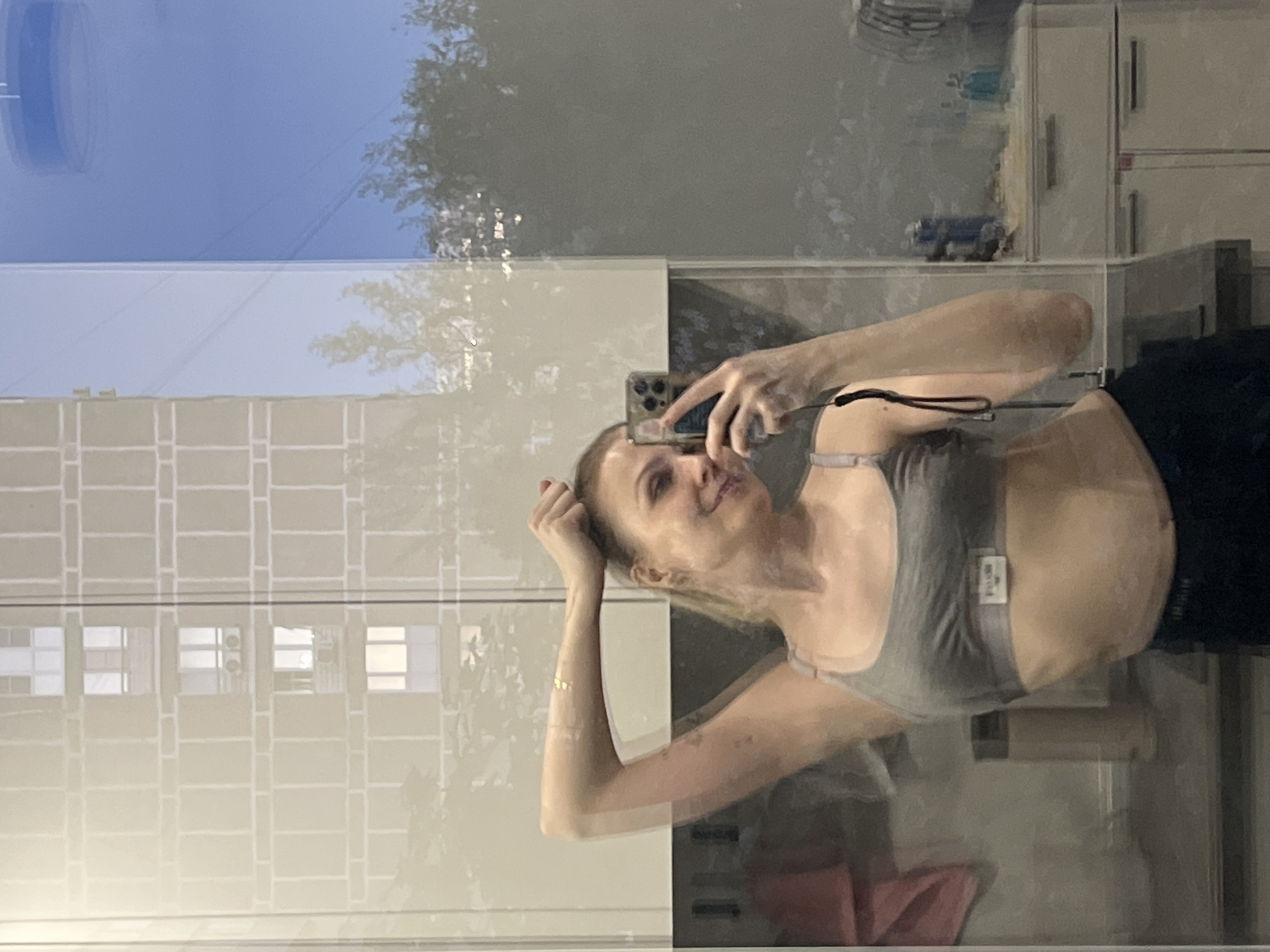 A mirror selfie captures a woman in a gray sports bra and black bottoms. The reflection shows a building with a grid-like pattern and glimpses of trees and sky. The background includes bathroom fixtures, with bottles, containers and drawers, with an additional towel on a rack. It is not possible to determine the specific location of Khimki, Russia from this image.
