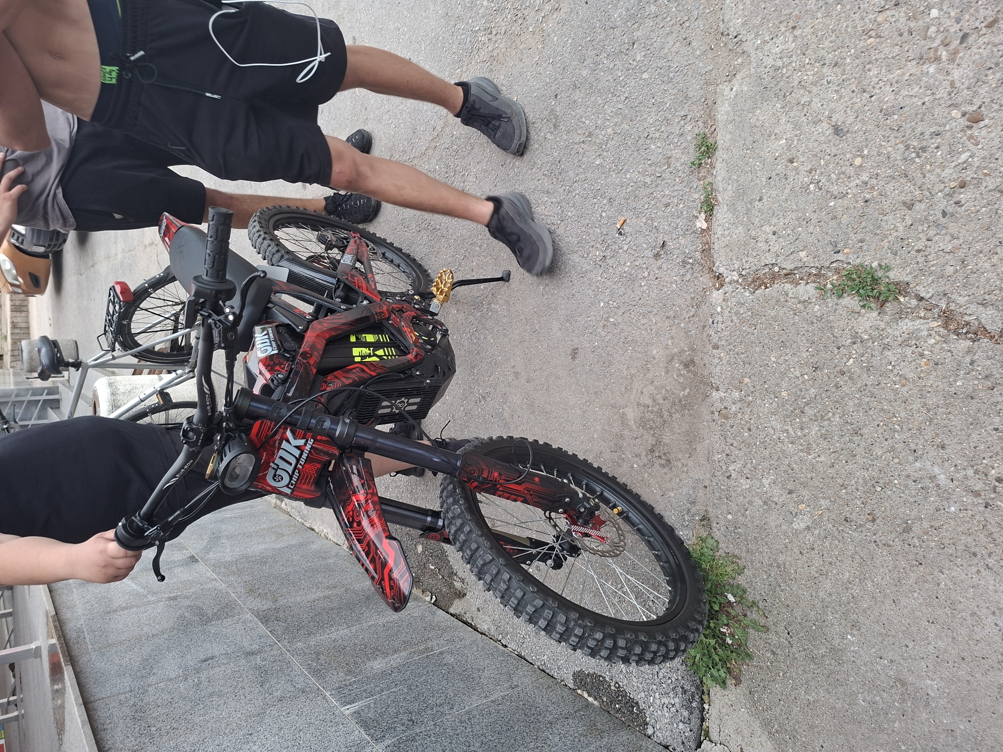 An outdoor scene in Basarbovo, Bulgaria, features three individuals positioned on cracked concrete pavement next to a grey, tiled building wall. Central to the image is a customized electric Sur-Ron dirt bike, predominantly black with striking red and black circuit board-style graphic detailing on its fenders, frame, and battery casing, alongside "GUK CHIP TUNING" branding. The bike is equipped with knobby off-road tires and a gold-colored kickstand.
On the far left, a person, wearing a black short-sleeved top and a black skirt, holds the left handlebar of the electric bike with their right hand. To the right of the bike, a second person is partially visible, wearing a grey t-shirt and black shorts, standing with their right hand on their hip. A dark-framed conventional bicycle with a rear rack is visible behind this individual. Further to the right, a third person is shown bare-chested, wearing black athletic shorts with a white drawstring and white wired earphones hanging from their left pocket. All three individuals are wearing black athletic shoes.
In the upper background, a gold-colored car is partially visible. The concrete pavement shows several cracks through which small patches of green vegetation are growing, and a discarded cigarette butt lies near the bike's kickstand. The scene is illuminated by bright daylight.