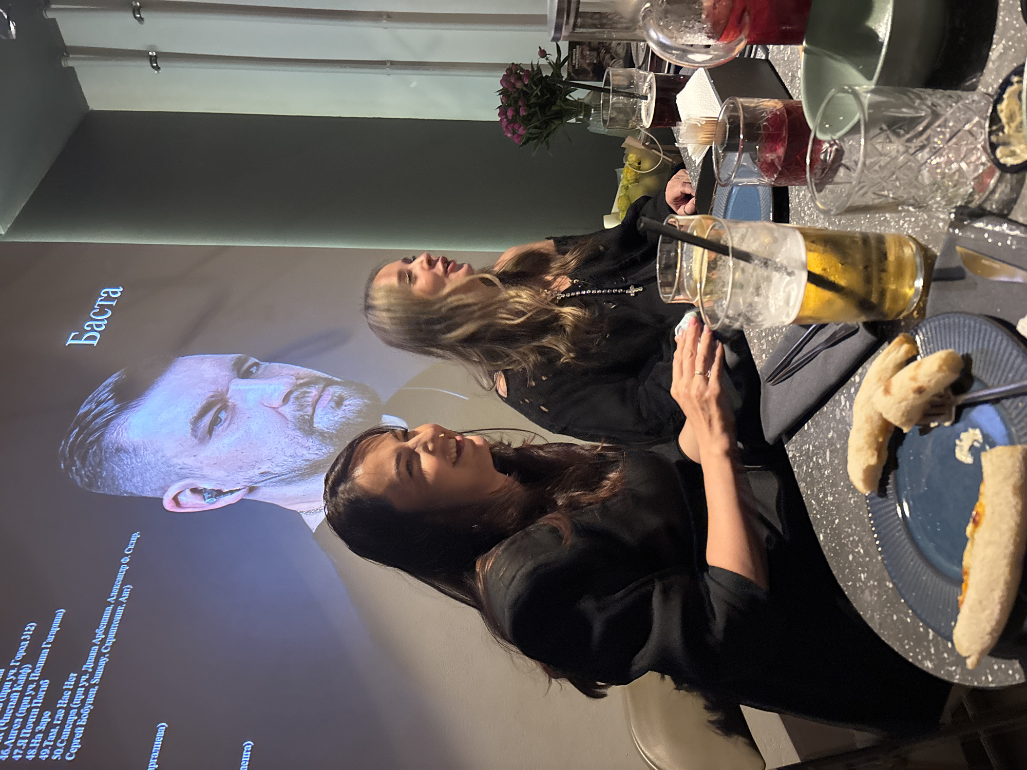 This image depicts an indoor scene in Chaikovsky, Russia, likely during the evening, possibly in a restaurant or an event venue. Two women are seated at a dark, speckled table, engaging in conversation or observing a large projection on the wall to their left. The woman in the foreground, with dark hair and wearing a black top, smiles while looking towards the projector. The second woman, with lighter, wavy hair and a dark top with a beaded necklace, is seated further back, looking towards the table with an open-mouthed expression suggesting laughter or speech.
On the table, various objects are present: several glasses containing drinks of different colors (some with ice and straws), a small vase with purple flowers, and a blue plate holding partially eaten food items resembling flatbread or pizza. The women's hands are visible, with one showing a ring.
The large projection on the wall displays a man's face, identified by the Cyrillic text "Баста" (Basta) next to him. Below his image, a numbered list of song titles and featured artists is visible. Notable text includes: "46. Ангел (при уч. Полина Гагарина)", "47. Я Почти Погиб", "48. На Заре", "49. Там, где Нас Нет", and "50. Сансара (при уч. Диана Арбенина, Александр Ф. Скляр, Сергей Бобунец, Sunsay, Скриптонит, Ант)". Other visible text fragments include "(Чистый Кайф)" and "(при уч. Город 312)". This suggests the projection is either a playlist from a concert, a music video, or an album presentation related to the artist Basta. The lighting is dim, emphasizing the glow from the projector screen.