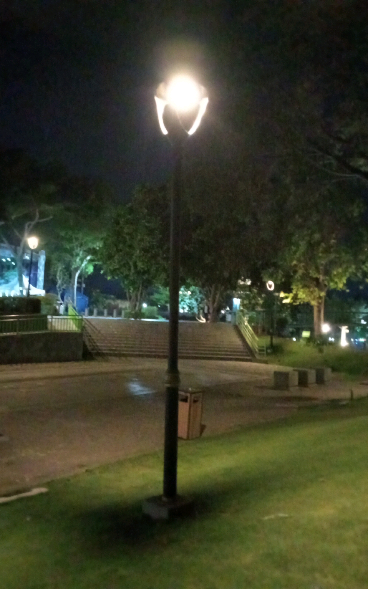 The image depicts a park scene at night in Ngasem, Indonesia. A tall, black lamp post with a lit, globe-shaped fixture stands prominently in the foreground, casting a bright light and a shadow onto the grass. To the left of the lamp post, a trash can is visible. Behind the lamp post, a paved pathway leads to a set of stone stairs. Trees with lush green foliage form the backdrop, illuminated by various lights. In the distance, a railing and what appears to be a structure with lights are partially visible. The ground is a mix of grass and paved areas, with some damp patches indicating recent rain. The overall atmosphere is dark and serene, characteristic of a park after dark.