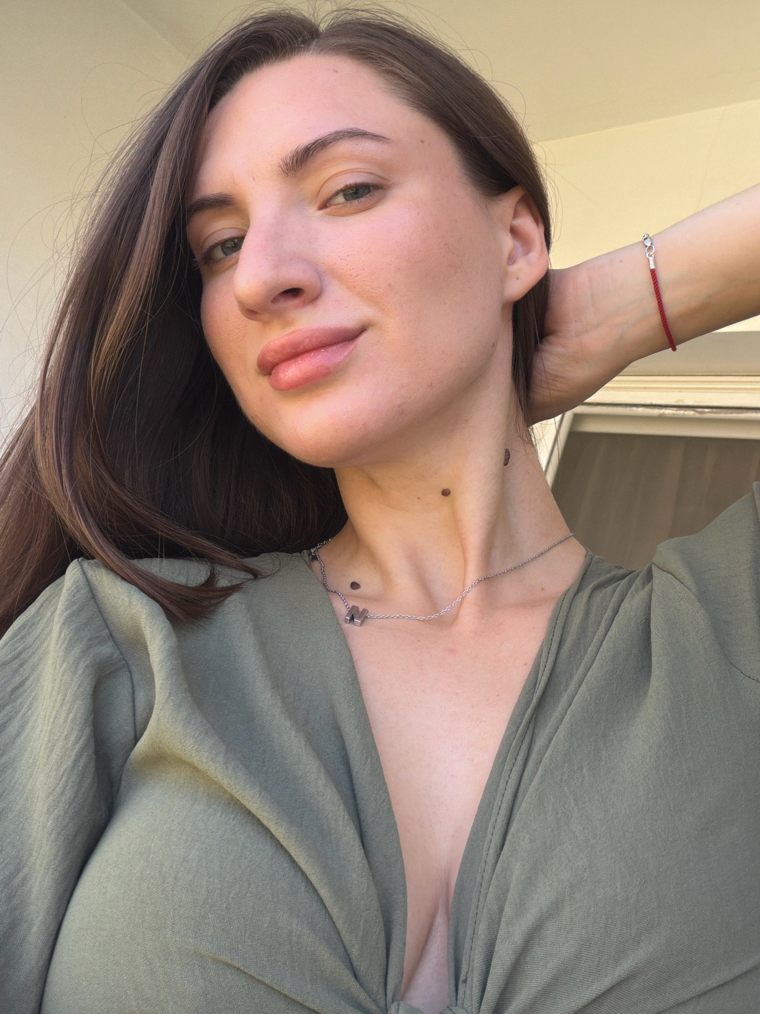 A young woman with long, dark brown hair, fair skin, and light eyes is looking directly at the camera. She is wearing a low-cut, olive-green top. She has a mole on her neck and another on her jawline. A thin silver necklace with a butterfly pendant is visible. On her left wrist, she wears a thin red string bracelet. Her right arm is bent and her hand is behind her head. The background appears to be outdoors, with a pale yellow wall or ceiling visible and a slightly out-of-focus window or door frame. There is no indication of the city of Sharm El Sheikh, Egypt.