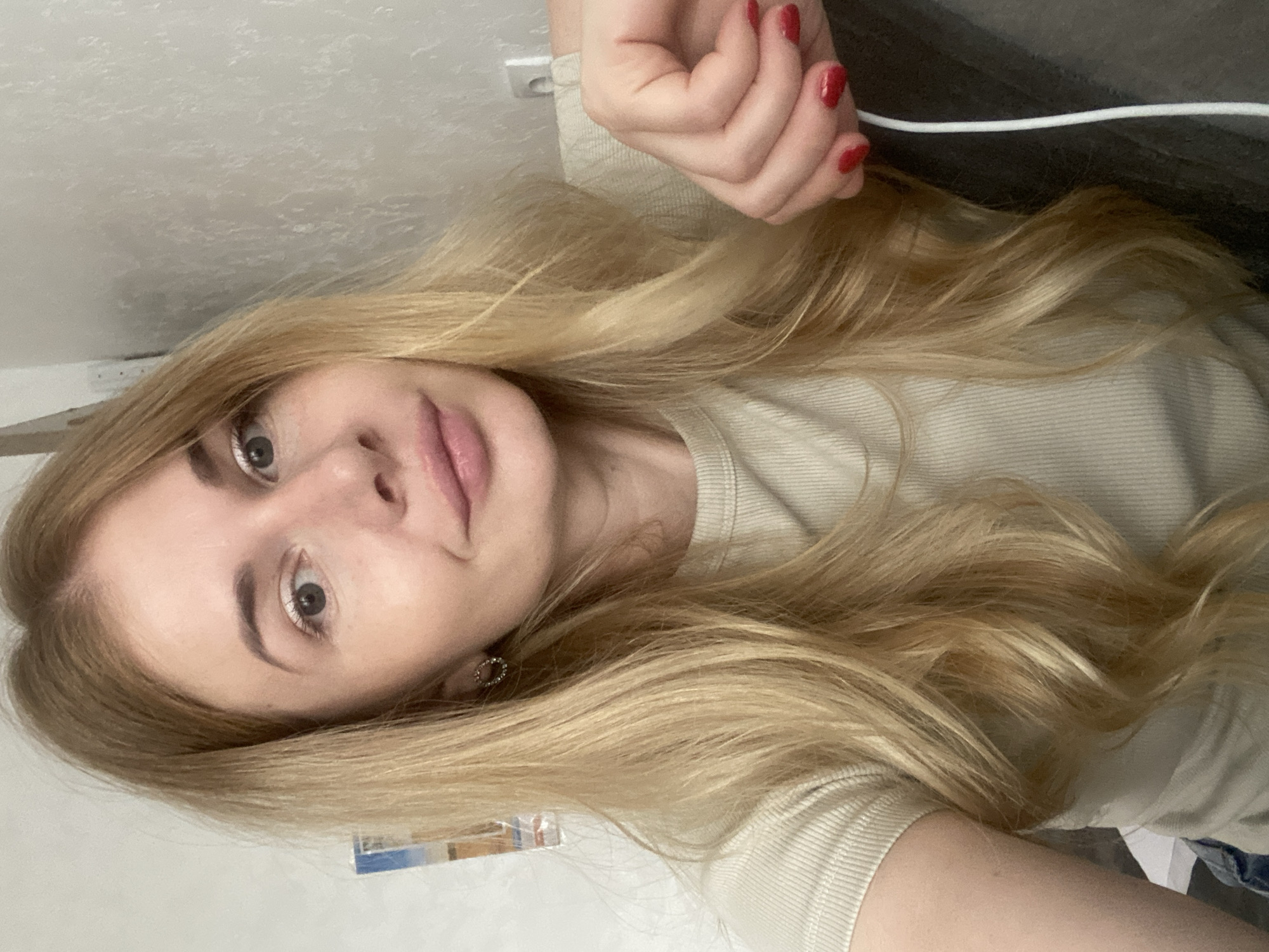 The image is a close-up, selfie-style shot of a young, fair-skinned woman with long, blonde, wavy hair. She has light-colored eyes, fair skin with a few freckles, and light pink lipstick. She is wearing a light-colored, ribbed t-shirt and has a silver, oval-shaped earring in her visible ear. Her right hand, with bright red nail polish, rests on a dark surface, possibly a couch. A white cord is visible near her hand. The background is a plain, light-colored wall with some texture. There is a hint of a framed picture on the wall to the left of the woman. The overall lighting suggests an indoor setting. There is no indication of the location being in the city of Voronezh, Russia.