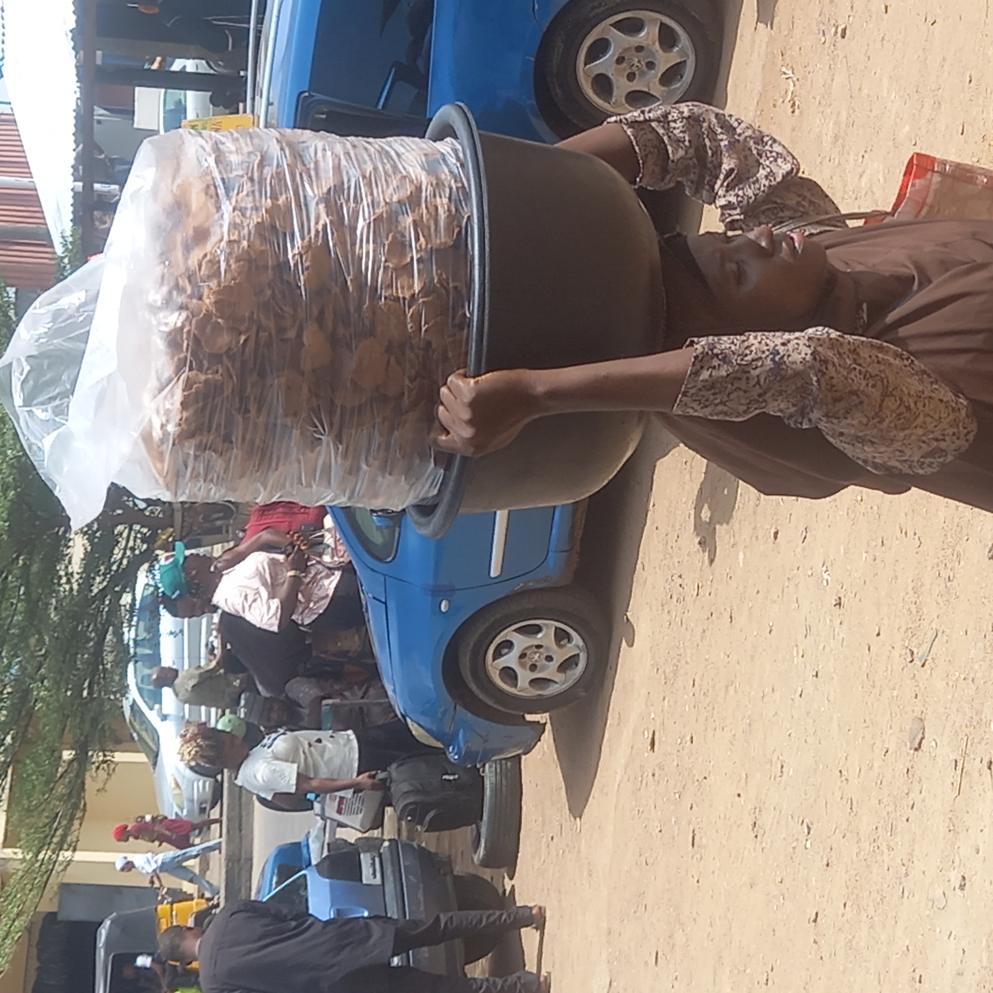 A woman carries a large bowl of bagged, flat, fried snacks on her head in Dadi Riba, Nigeria. She stands next to a blue car, presumably for selling. Other people and vehicles are in the background on a sandy street.