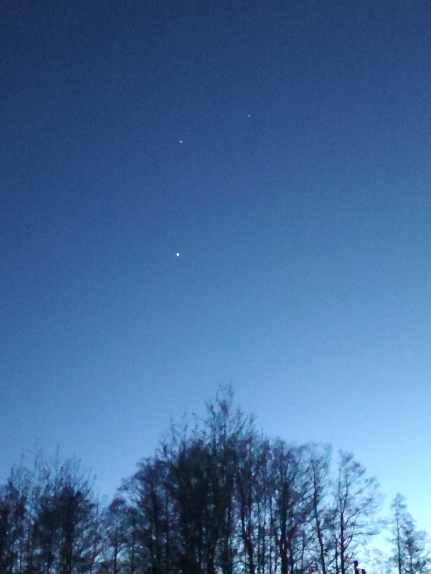 The image shows a dark blue sky with three bright spots, possibly stars or planets, in the upper portion. Silhouetted against the sky are the bare branches of trees. The location is likely Chaikovsky, Russia, based on context. There are no visible people, discernible subjects beyond the sky and trees, or actions taking place. The environment appears to be twilight or night.