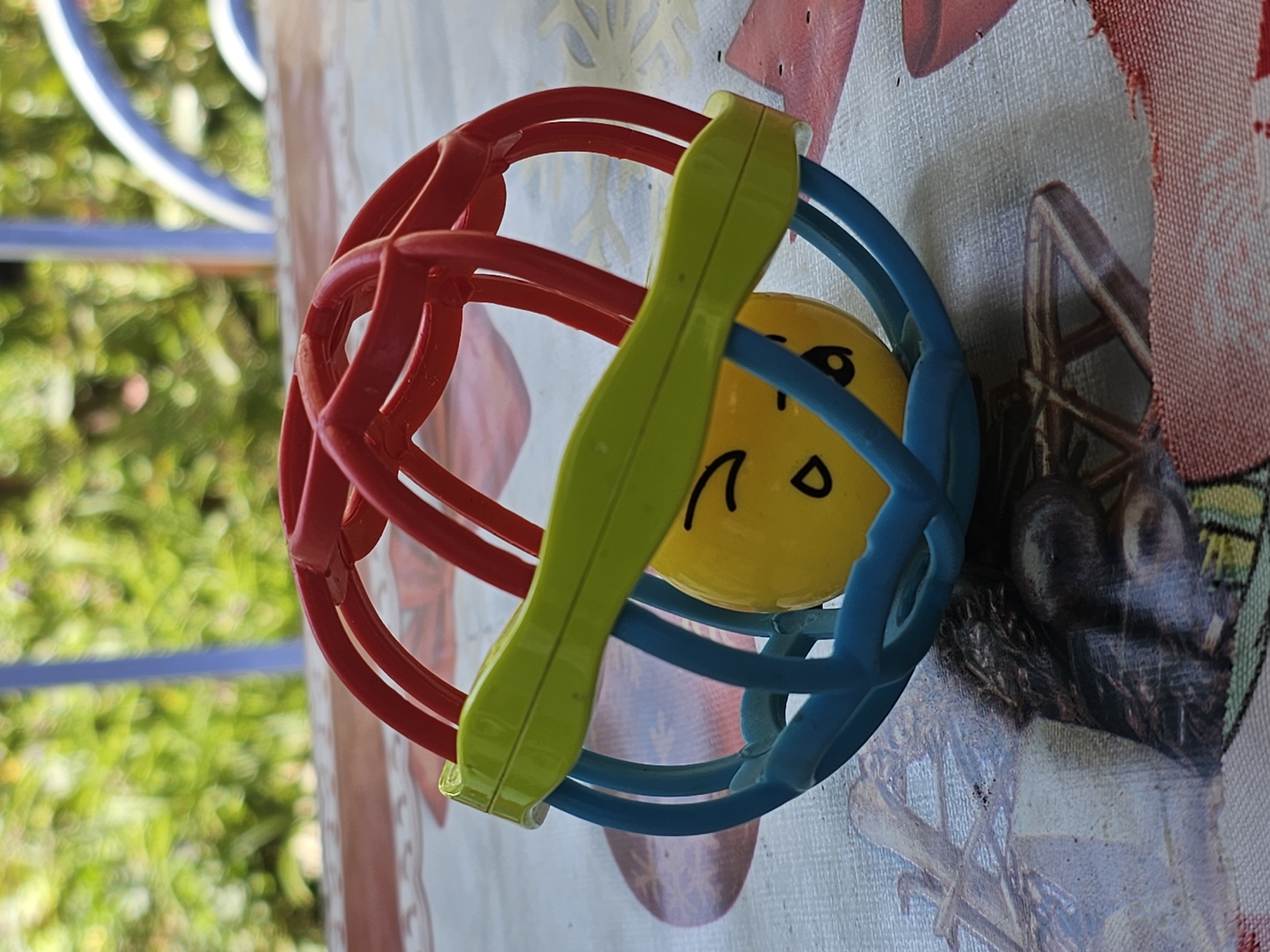 A baby rattle with a yellow ball face and a cage of red, blue, and green plastic bars rests on a patterned tablecloth. The background shows blurred green foliage and parts of a blue structure, suggesting an outdoor setting. The scene is set in Özgön, Kyrgyzstan.