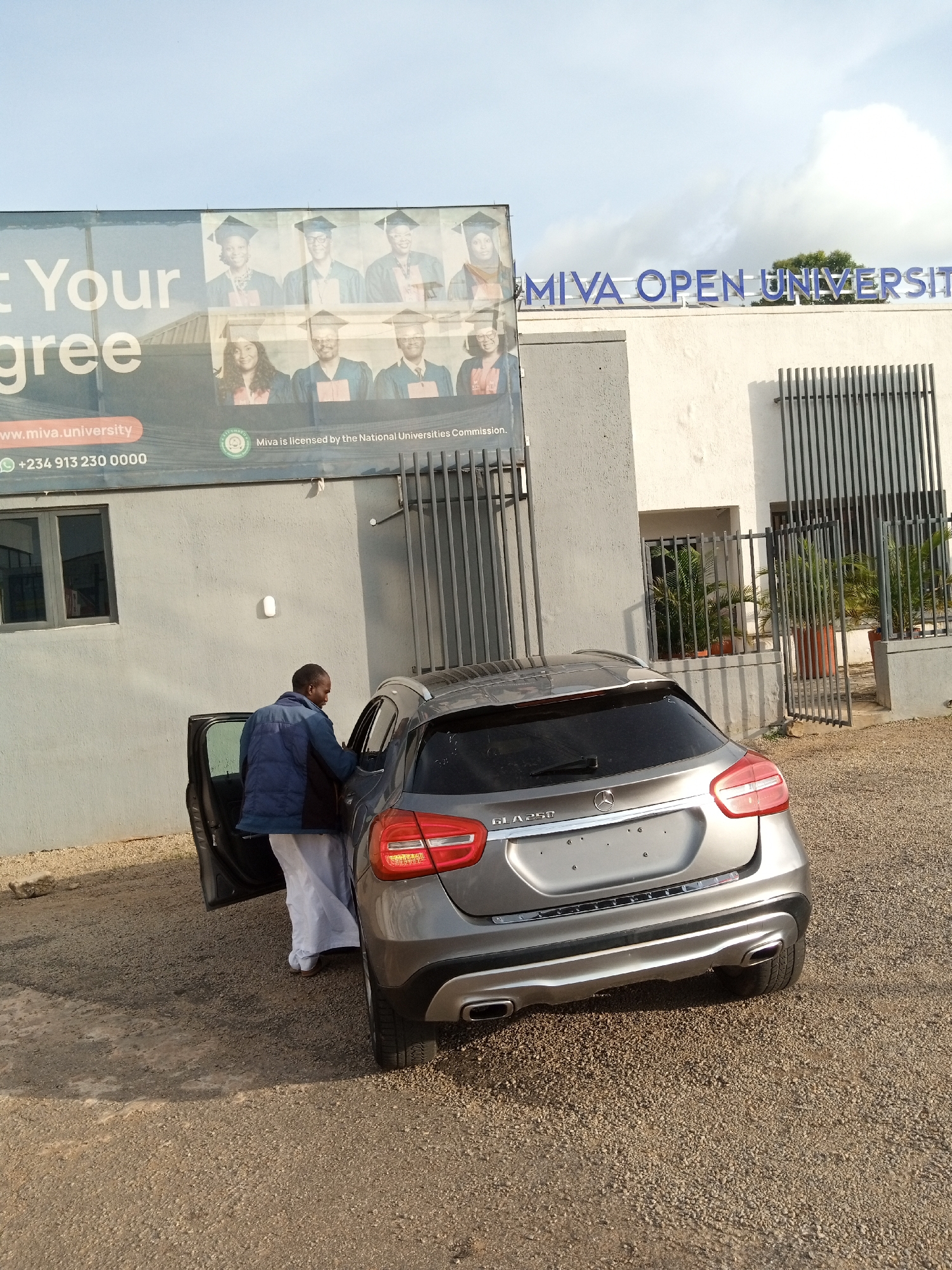 The image depicts the front of MIVA OPEN UNIVERSITY in Jos, Nigeria, with a billboard advertising their programs. A man, dressed in a blue jacket and white pants, is seen entering the driver's side of a silver Mercedes-Benz GLA 250, which is parked on a gravel lot in front of the building. The university building is light-colored with barred windows and signage. The billboard features images of graduating students and contact information for the university. The sky is partly cloudy.