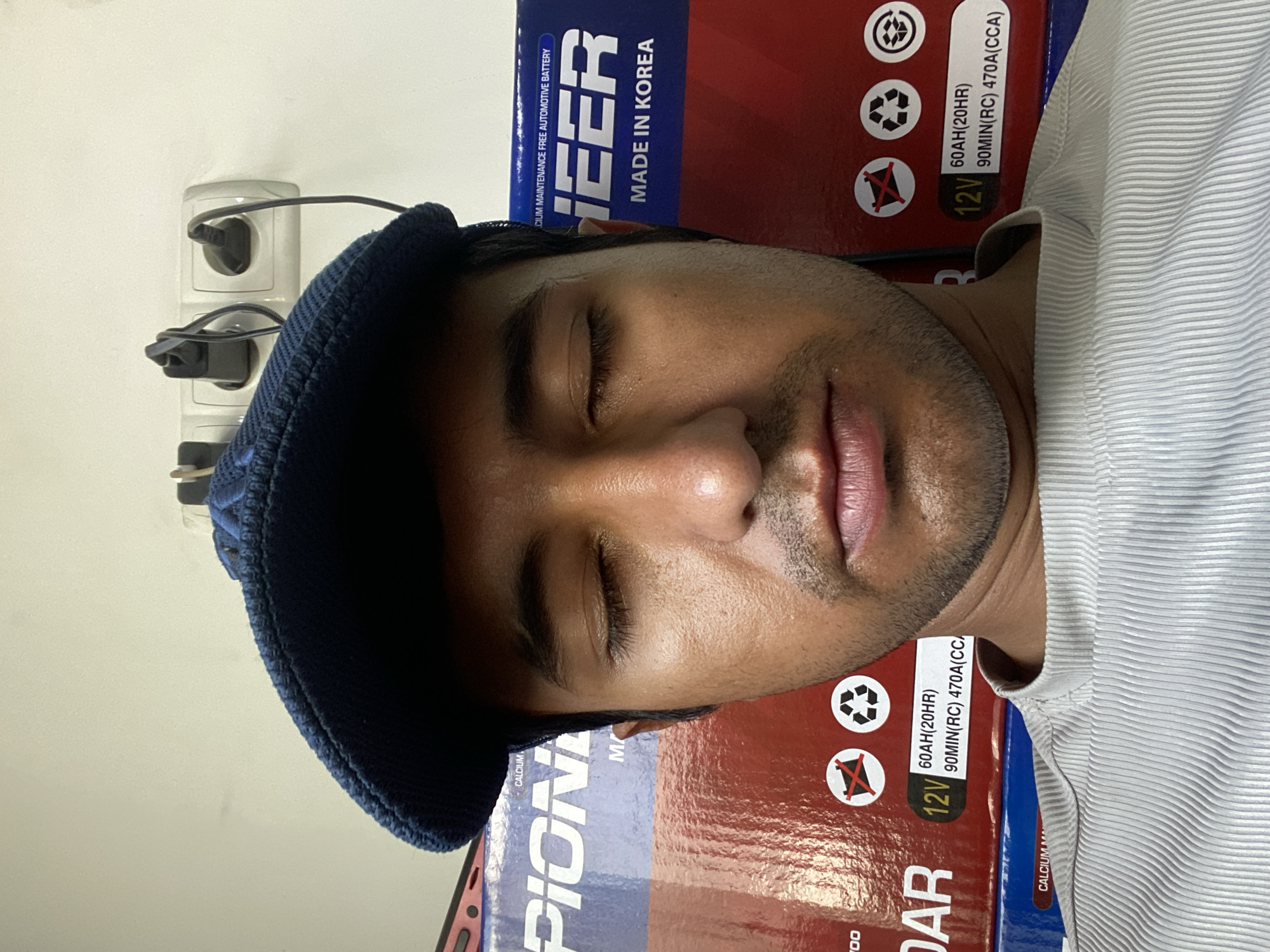 A man is lying on top of two battery boxes, sleeping. The battery boxes are red and blue and have the brand name "Pioneer" printed on them. The man is wearing a blue knitted cap and a white striped shirt. There is a white wall behind him with an electrical outlet. The text on the battery boxes indicates "Made in Korea." There is no indication that this image was taken in Bukhara, Uzbekistan.