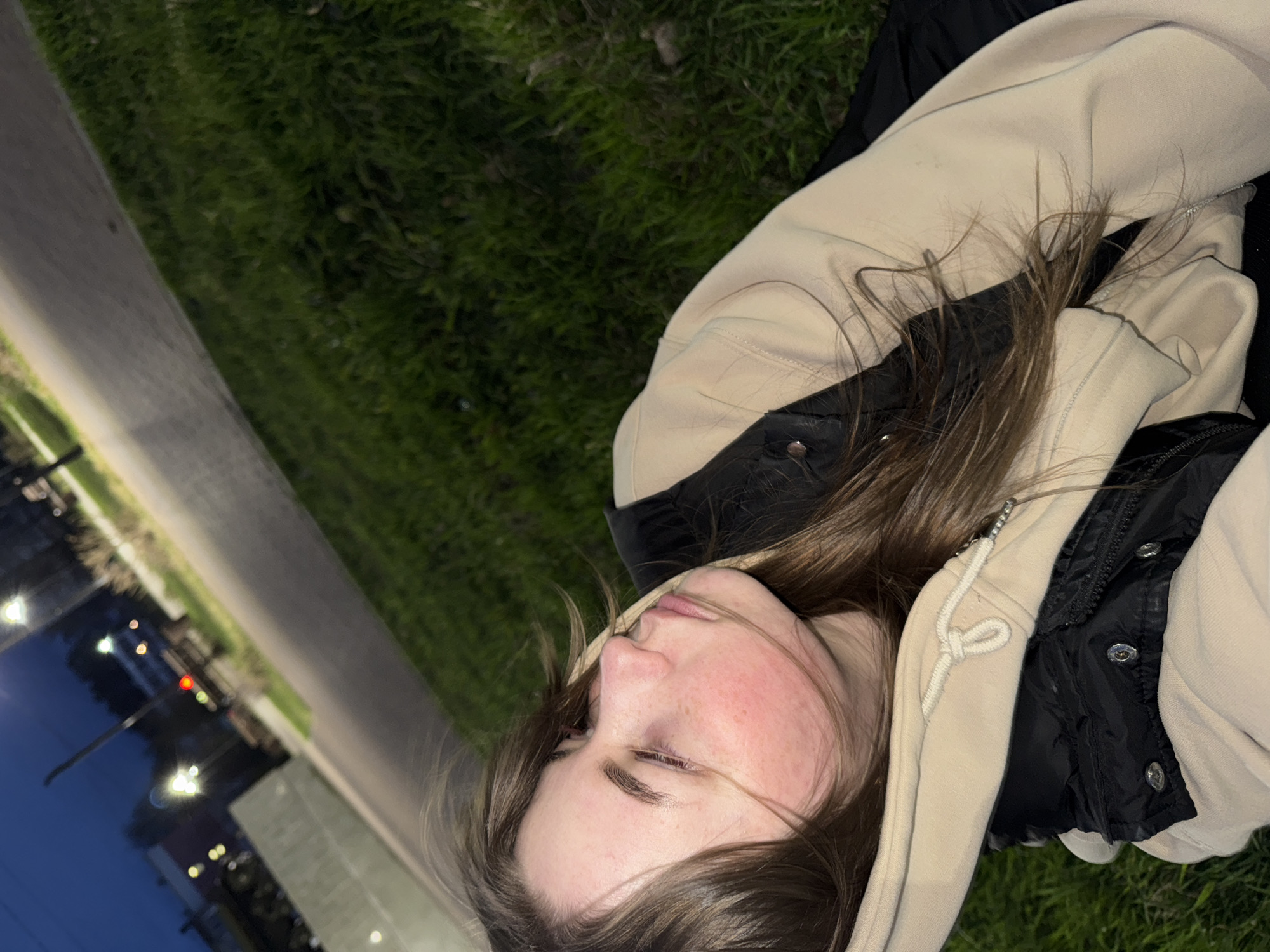 A close-up, rotated image captures a young person, likely female, lying down outdoors during the evening or night in Chaikovsky, Russia. The subject's head and upper torso are visible, angled as if resting on their back. They have long, dark brown hair, with some strands softly falling across their face. Their eyes are closed or cast downwards, conveying a relaxed or contemplative mood. The person exhibits a natural flush on their cheeks and visible freckles.
They are dressed in a light beige hooded sweatshirt layered over a black quilted or padded jacket, featuring a zipper and silver snaps.
The setting appears to be an outdoor urban or suburban area. Green grass is visible to the lower right of the subject, suggesting a lawn or park. A grey paved path or road runs along the left side of the frame. The background is blurred, showcasing a dark, evening sky dotted with numerous out-of-focus artificial lights, including bright white and one prominent red light, indicative of streetlights or distant building illumination. No text is visible in the image.