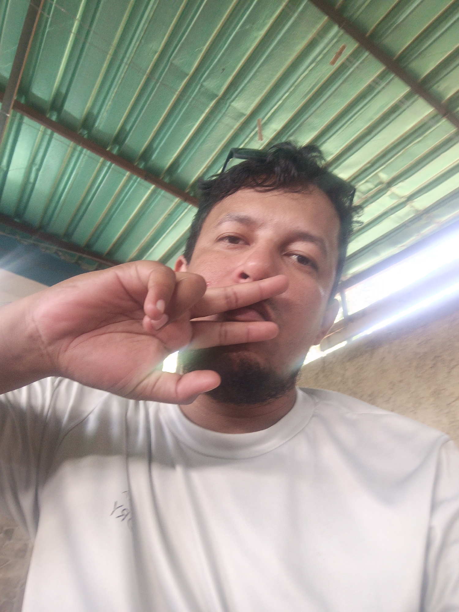 A man is captured in a close-up, low-angle selfie. He is wearing a white t-shirt. His left hand is raised, with his index and middle fingers extended in a 'peace' sign, positioned in front of his mouth. The man has dark hair and a beard. The background consists of a corrugated metal ceiling, painted green, with a brown metal beam visible. To the right of the man, a bright light source is emanating from what appears to be a window or opening, illuminating a textured wall. The scene suggests an outdoor or semi-outdoor setting, possibly a covered patio or a structure with a metal roof, typical of some areas in Juan Griego, Venezuela.