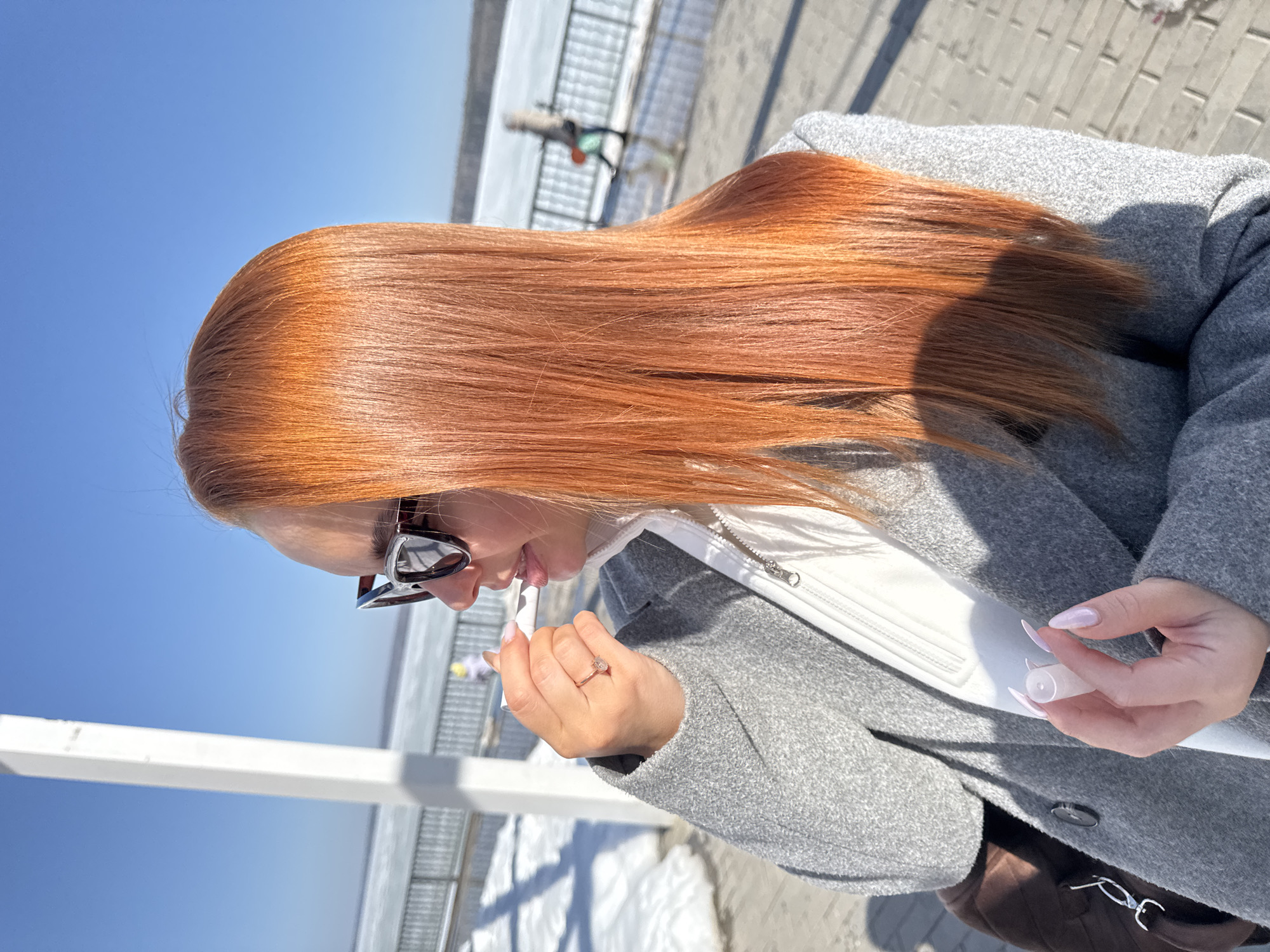 A woman with long, straight auburn hair and sunglasses is applying lip gloss on an outdoor boardwalk. She is wearing a grey wool coat over a white top. A brown purse is visible on her shoulder. The location is likely a boardwalk or promenade in the city of Chaikovsky, Russia, near an icy body of water with metal railings. The sky is clear and blue.