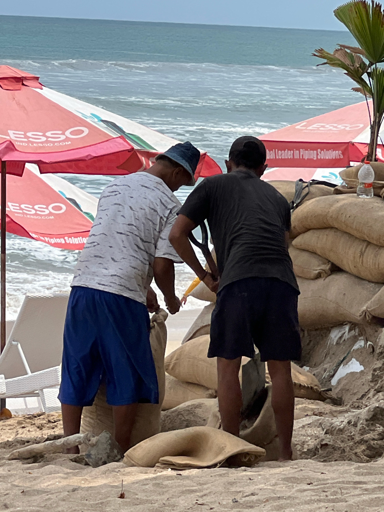 Two individuals are working on a sandy beach in Kuta, Indonesia, near the ocean. The water is visible in the background with waves. In the foreground, several large, sand-filled bags are stacked, forming a barrier. One person, wearing a blue hat and a patterned t-shirt with blue shorts, is kneeling and appears to be filling a sandbag with a shovel. The other person, wearing a black t-shirt and black shorts, is standing and holding a shovel, seemingly assisting with the task. Red umbrellas, branded with "LESSO," are positioned behind the individuals, suggesting a commercial area. A palm frond is visible to the right, and a plastic bottle of water sits atop the sandbag barrier. The overall scene depicts an effort to reinforce the beach area, possibly due to coastal erosion or impending weather.