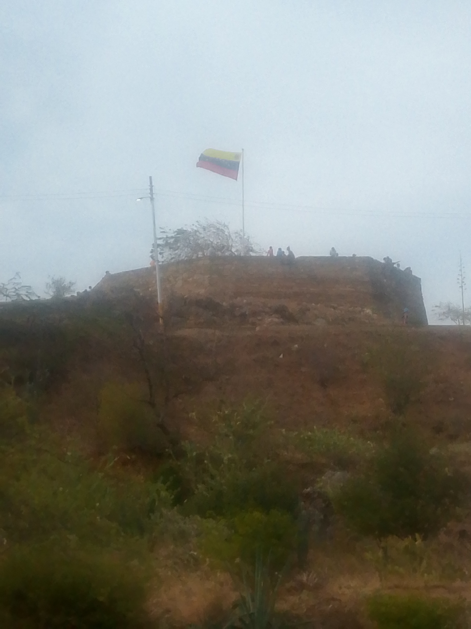 The image depicts a hill or fortification in Juan Griego, Venezuela, with the Venezuelan flag flying atop it. Several people are present on the top of the hill. The foreground features vegetation, suggesting the hill is naturally integrated into the landscape. A lamppost stands near the base of the flagpole. The sky is overcast.