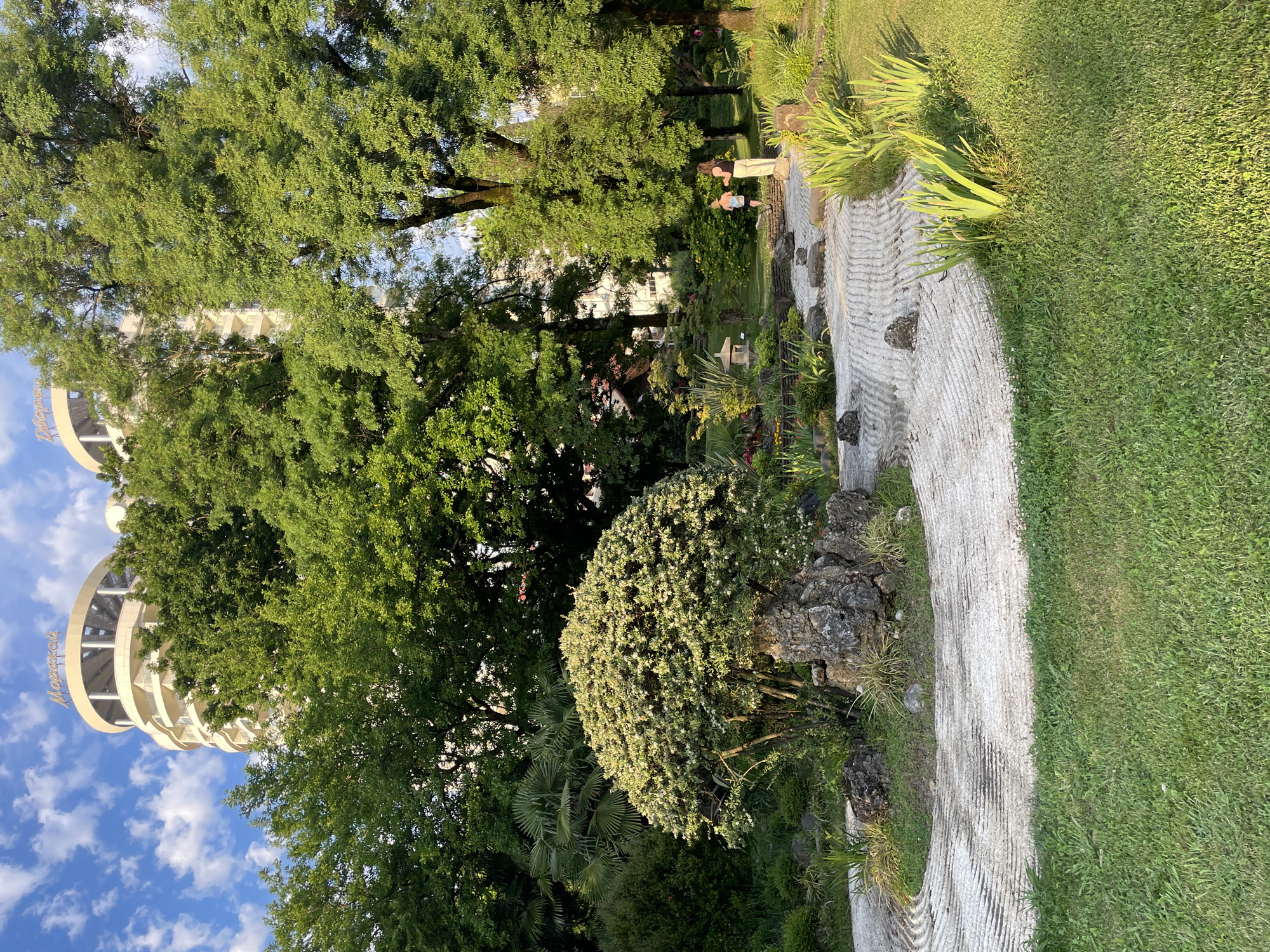 The image depicts a serene Japanese garden in Sochi, Russia, likely part of the Dendrarium Botanical Garden or a similar park. In the background, a tall, modern building with a rounded facade and the word "Морской" (Morskoy - meaning "Maritime") visible on top, suggests its proximity to the coast. The garden features meticulously raked white gravel, creating wavy patterns that symbolize water. Large rocks are strategically placed, with one particularly prominent rock serving as a base for a rounded, bushy tree. Smaller rocks and lush green vegetation, including palm trees and flowering plants, populate the garden. Two individuals, a woman and a child, are visible in the middle ground, suggesting a peaceful, leisurely atmosphere. The overall scene is bathed in soft sunlight, indicating a clear day.