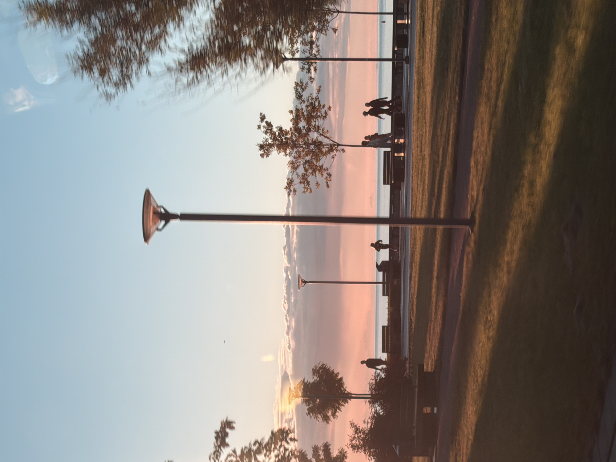 The image depicts a sunset scene in Tallinn, Estonia, likely along the coastline or a waterfront promenade. The sky is filled with soft hues of pink and orange, transitioning to a pale blue overhead, with scattered clouds. In the foreground, a grassy park area is illuminated by long shadows cast by the setting sun and a lamppost. A paved path runs through the park, bordered by benches and trees.
Further back, a waterfront promenade is visible with several lampposts and benches. Silhouettes of people can be seen walking and standing along the promenade. One individual is on a scooter. The sea is visible in the background, appearing calm. The overall atmosphere is tranquil and serene.