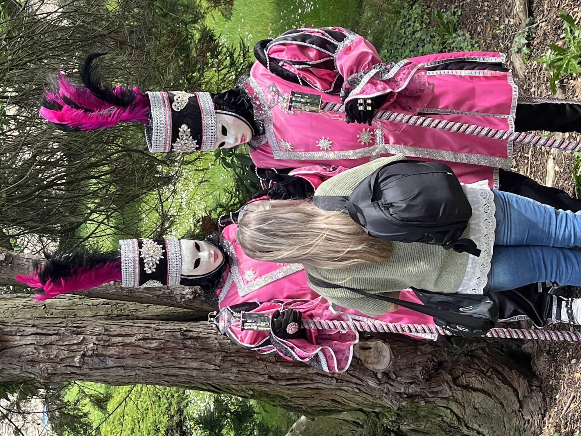 The image depicts two individuals dressed in elaborate, colorful costumes reminiscent of a Venetian carnival, positioned alongside a tree in what appears to be an outdoor setting in Saint-Galmier, France. The costumed figures wear pink and black outfits with ornate silver detailing, masks, and feathered headpieces. A woman wearing a green sweater and blue jeans stands slightly behind one of the figures, obscuring the lower portion of the costume. She has a black backpack and tote bag. The background includes foliage and greenery, suggesting a park or garden setting. The photo is rotated 90 degrees clockwise.