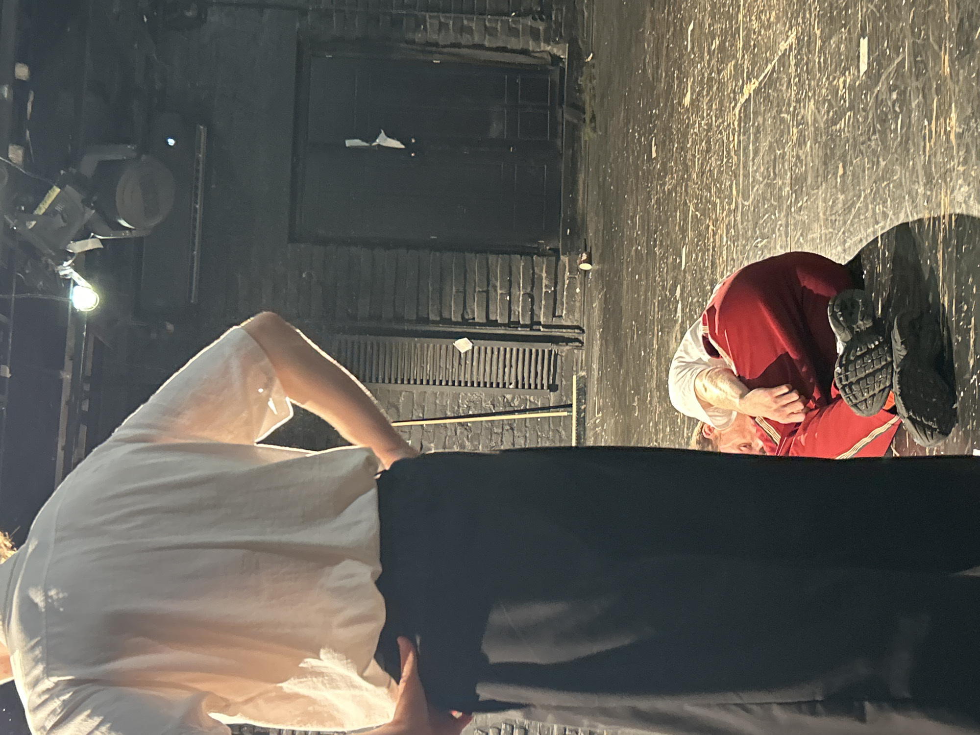 Two people are on a stage with a brick wall in the background. A person in the foreground is standing with their back to the camera, wearing a white shirt and black pants. Another person is lying on the floor in the background, wearing a white shirt and red pants, with their eyes closed. The stage floor is made of wood and appears worn. Stage lights are visible overhead.