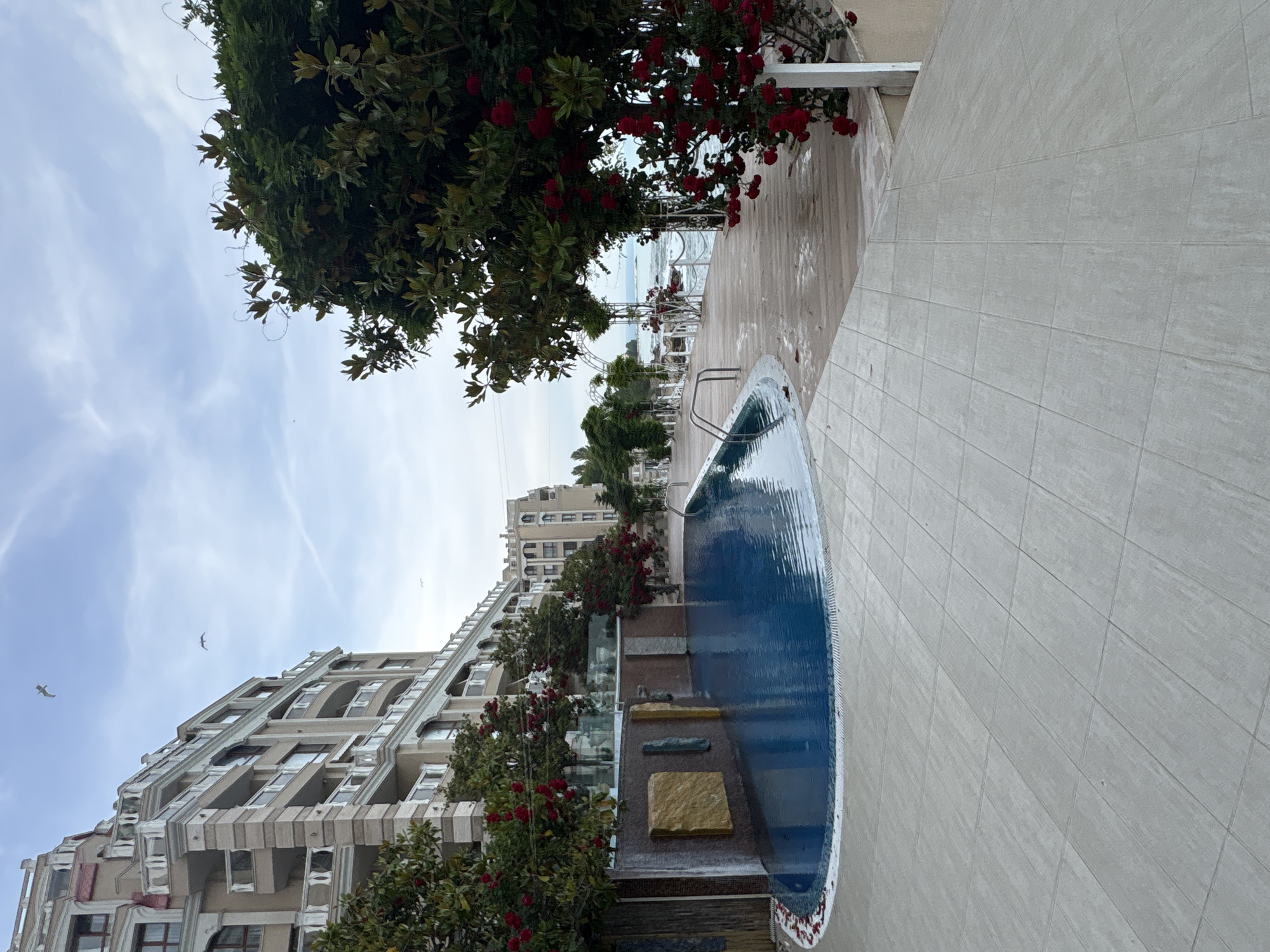 The image captures a serene outdoor scene at what appears to be a coastal resort or hotel. The perspective is slightly elevated, looking towards a body of water under a partly cloudy sky.
In the foreground, a light-colored tiled patio stretches across the bottom and right side of the frame. On the right, a blue swimming pool with a curved shape and visible steps is filled with rippling water, reflecting the sky. Along the left edge of the patio and bordering the pool are lush green trees and bushes adorned with numerous vibrant red flowers, possibly roses.
Behind the foliage, a large, multi-story building dominates the left side of the midground. It features intricate architectural details, including multiple balconies with railings and arched windows, suggesting a resort or residential complex. Several birds, likely seagulls, are seen flying in the pale blue sky above the building.
In the background, beyond the trees and pool area, a calm expanse of water, presumably the sea or ocean, meets the horizon. Some white structures, possibly sun loungers or pergolas, are visible near the water's edge.
The scene is tranquil with no specific human activity depicted. The time of day appears to be daytime, with soft, diffused natural light indicating either morning or late afternoon. The weather is partly cloudy. The overall impression is one of peaceful relaxation in a picturesque seaside setting. No text is visible in the image.