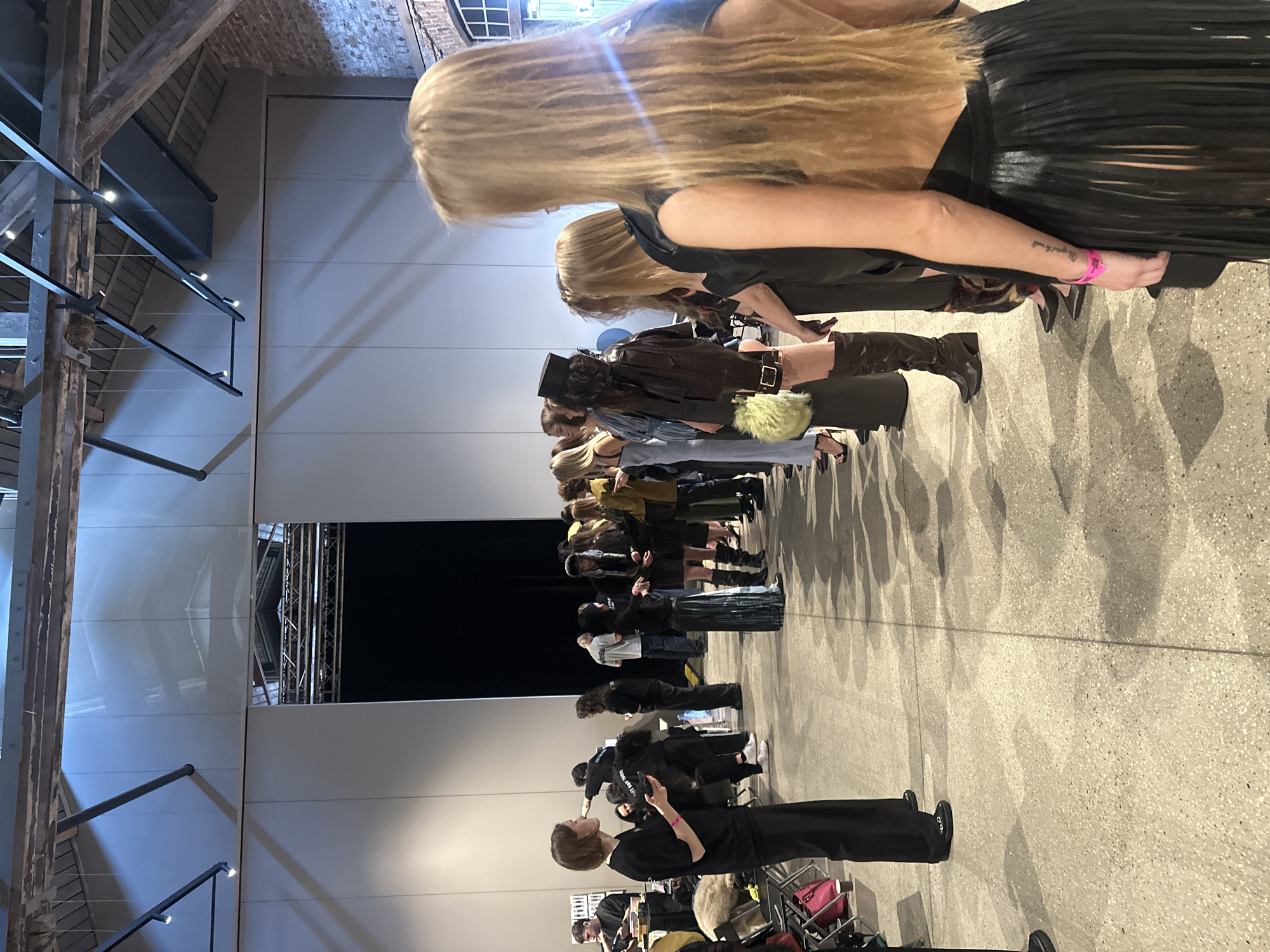 The image captures an indoor scene, likely a backstage or pre-show area at a fashion event in Riga, Latvia. A long line of people, predominantly women, stretches into the background against a plain light-colored wall.
In the foreground, a person with short brown hair, dressed in a black top and trousers, is seen holding a phone and facing towards the line, seemingly overseeing or interacting with the participants. Other crew members or staff, also mostly in black attire, are visible in the foreground and mid-ground, some seated or attending to items.
The line is comprised of models, many with long, flowing light-colored hair, dressed in various fashionable ensembles. Outfits include a black pleated skirt, a dark leather jacket, black boots, and other stylish clothing. A prominent detail is a bright, fluffy green or yellow handbag worn by a model in the mid-ground.
The venue features industrial-style architecture, with exposed wooden beams, brick walls, and modern metal light fixtures visible in the upper parts of the room. The floor is a light, speckled concrete or terrazzo, reflecting the overhead lighting and casting long shadows of the people. In the far background, a large, dark rectangular opening suggests a stage entrance or a dedicated presentation area.
Visible text includes a pink wristband on the right arm of a model in the foreground, below which a tattoo reads "My heart is full". On a black T-shirt worn by a person in the mid-ground, the partial text "WE AIN'T NO..." is discernible. Further back, on other black tops, "ON ME." and "ONLY THE..." can be seen. The overall mood appears to be one of preparation and focused anticipation.