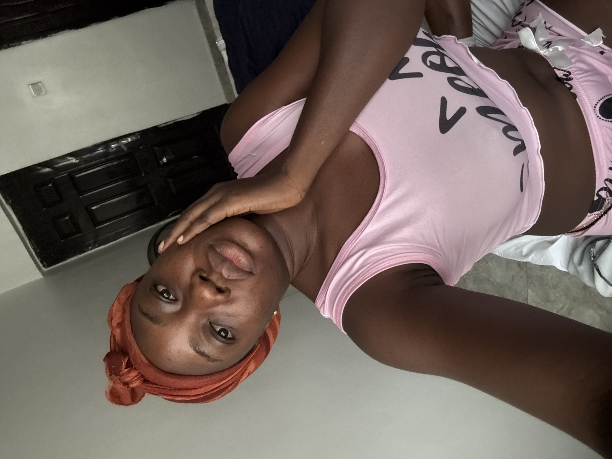 An indoor portrait shows a young adult Black woman lying down, looking directly at the camera. She wears a light pink sleeveless top with stylized black text, and matching pink patterned shorts with a white bow. Her hair is covered by an orange or rust-colored headwrap. Her right hand is gently placed near her face, which has a calm and neutral expression, with her lips slightly parted. The background features a light grey wall with a small, square, metallic fixture, possibly an electrical outlet. Below this, a dark grey or black panel with rectangular indentations is visible, suggesting a piece of furniture or a bed frame. White fabric, likely bedding, is also present on the right side of the frame. The scene conveys a relaxed atmosphere in a personal space, located in Iddo Tudunwada, Nigeria.