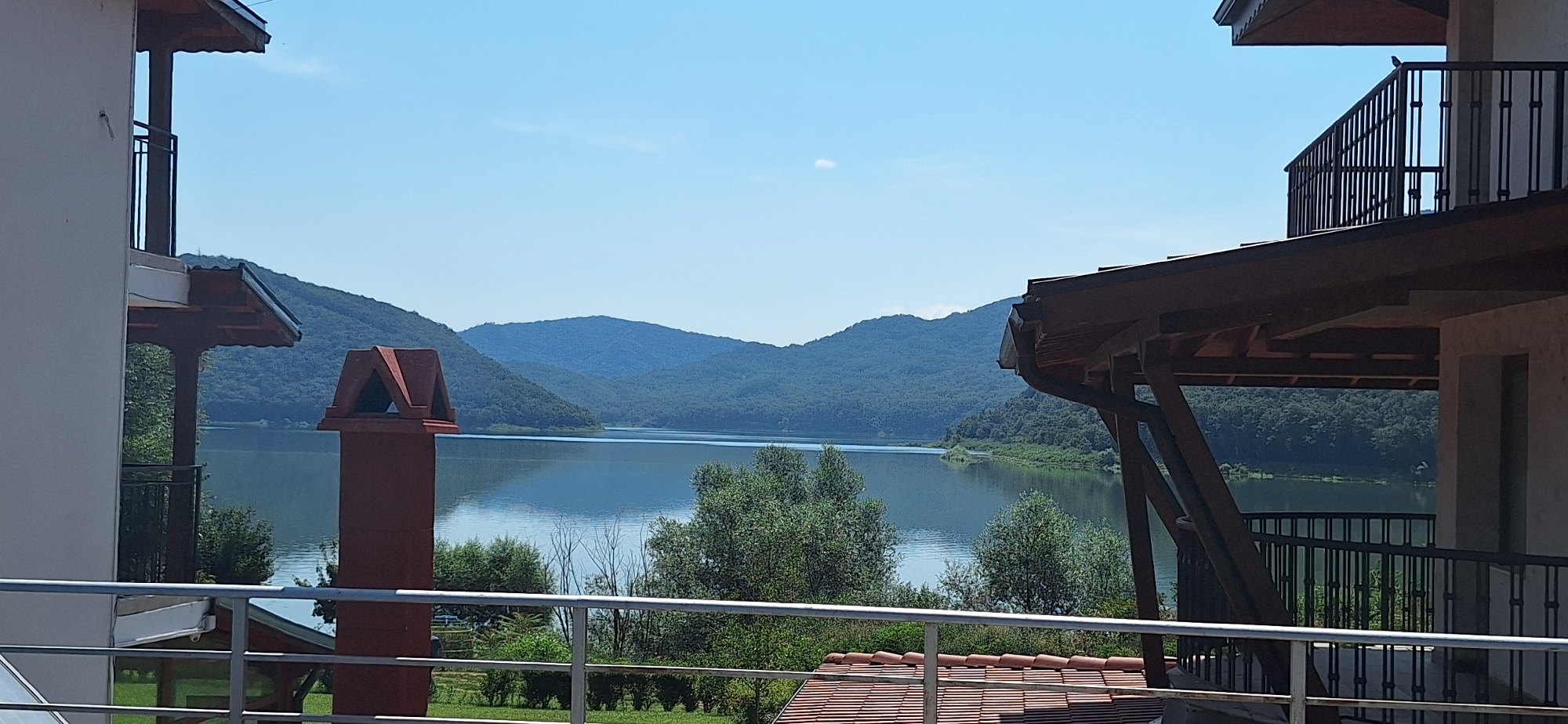 The image captures a scenic view of the Asparuhovo dam reservoir, located near the city of Asparuhovo, Bulgaria. The perspective is from a balcony or elevated platform of a building, looking out towards the water and surrounding hills. In the foreground, architectural elements of the building are visible, including a red brick chimney with a decorative cap, railings, and parts of the roof structure. The lake itself is calm, reflecting the clear blue sky and the lush green, tree-covered hills that encircle it. The day appears to be sunny and clear, with a few wispy clouds in the sky. The scene suggests a peaceful, natural setting with human habitation integrated into the landscape. There are no people or animals visible in the frame.