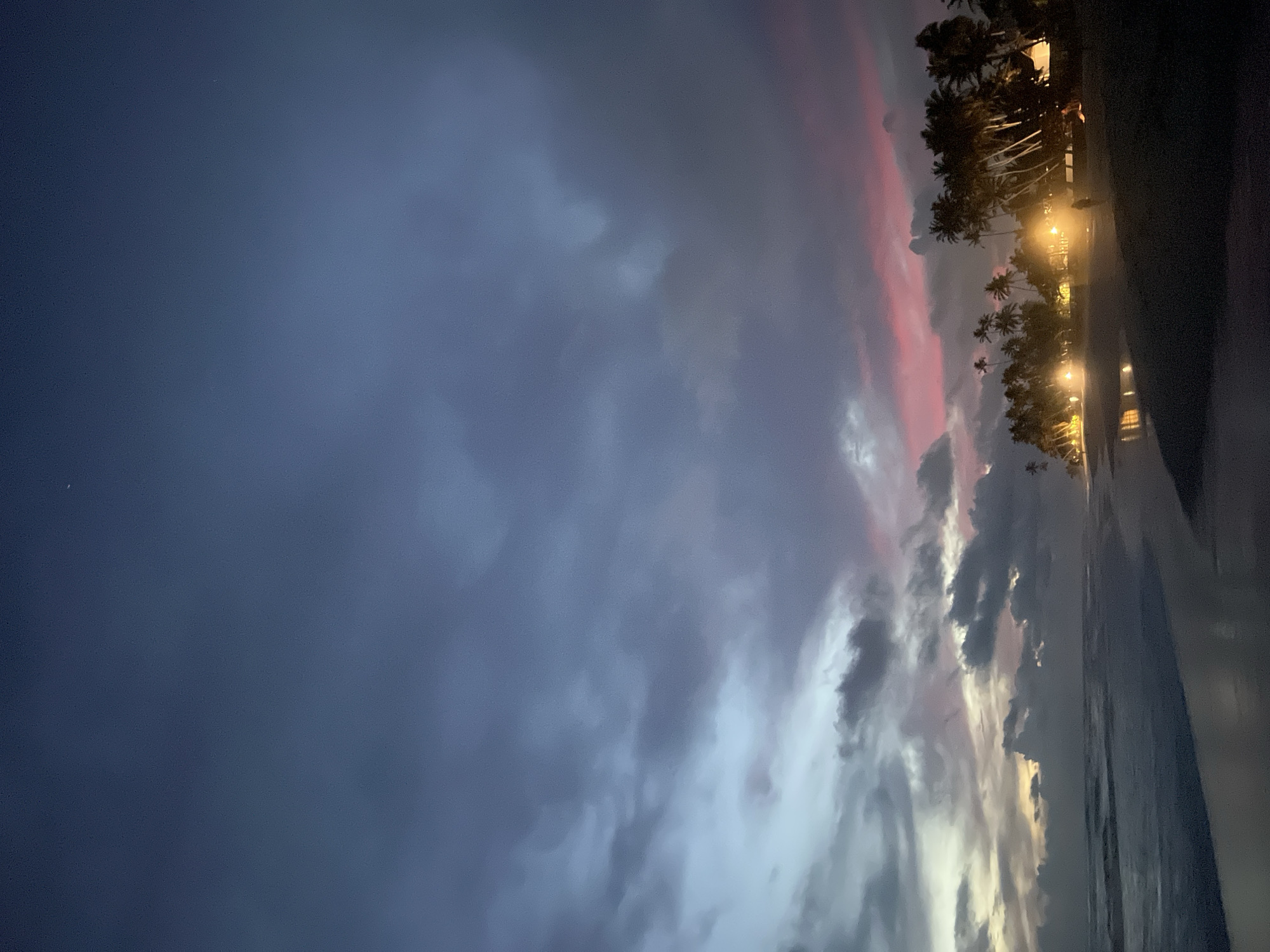 This vertical image depicts a tropical coastal scene at twilight or night, with the sky dominating the composition. The upper portion of the sky is a deep, dark blue-grey, gradually transitioning to lighter grey, white, and striking streaks of pink and red towards the right side. Dense, textured clouds are scattered across the sky, reflecting the fading light. A tiny, bright white dot, possibly a star or the moon, is visible in the upper left of the dark sky.
Along the right edge and bottom of the image, a line of tall palm trees stands in silhouette against the lighter sky, suggesting a beach or resort area. Below these trees, multiple bright artificial lights cast a warm glow, illuminating unseen structures or parts of the ground. The very bottom right shows a dark expanse, likely the ocean or a wet beach reflecting some light. The overall atmosphere is serene and dramatic, characteristic of a tropical evening. The location is Habaraduwa, Sri Lanka, which aligns with the tropical beach setting. There is no visible text in the image.