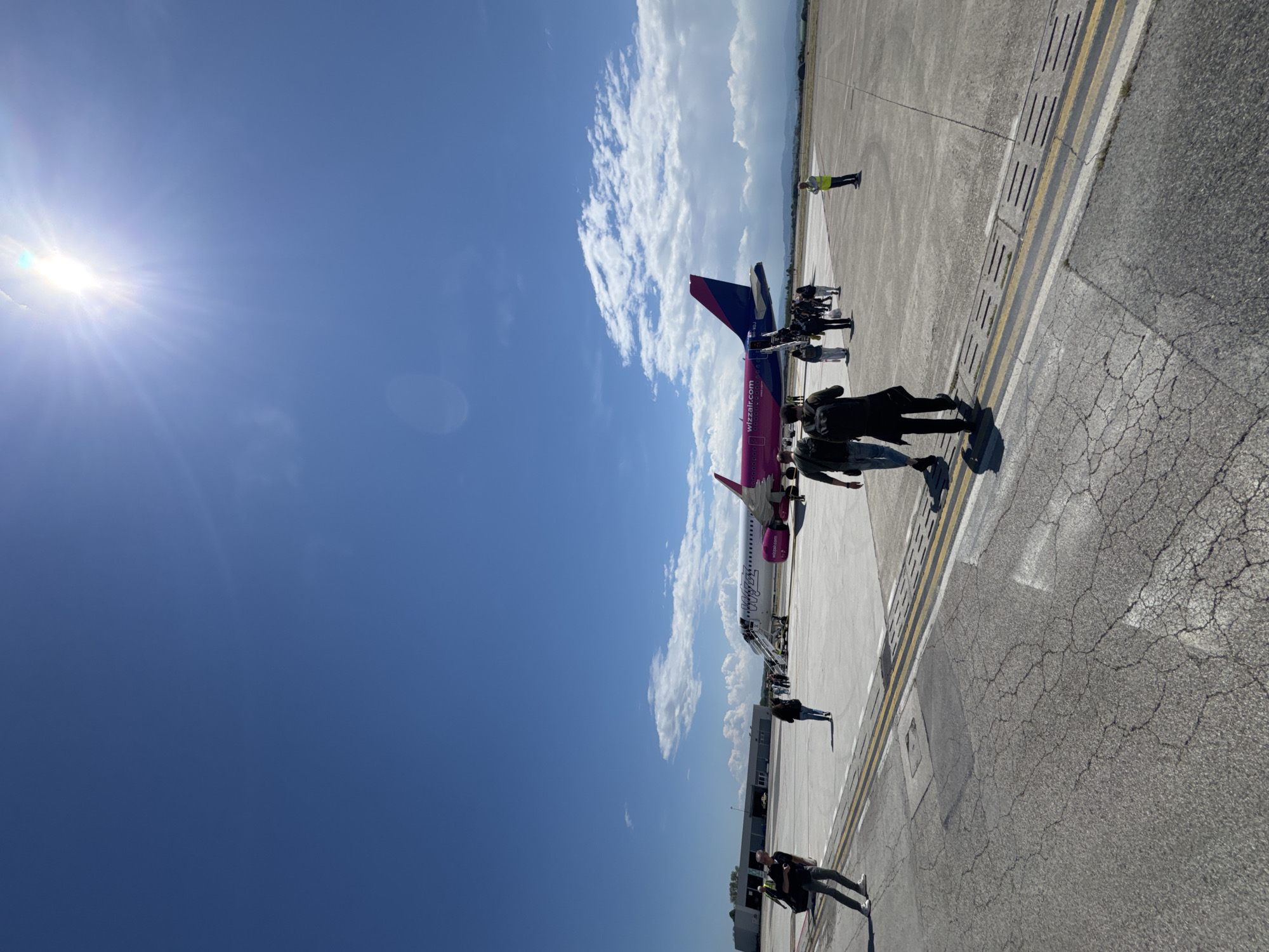 The image shows an airplane on an airport tarmac under a bright blue sky with scattered clouds. People are boarding or disembarking the plane. The airplane is painted in the colors of the airline Wizzair. The ground is paved with asphalt and features yellow markings. The image was likely taken in or around Rimini, Italy, as Wizzair operates flights to and from Rimini Federico Fellini International Airport.